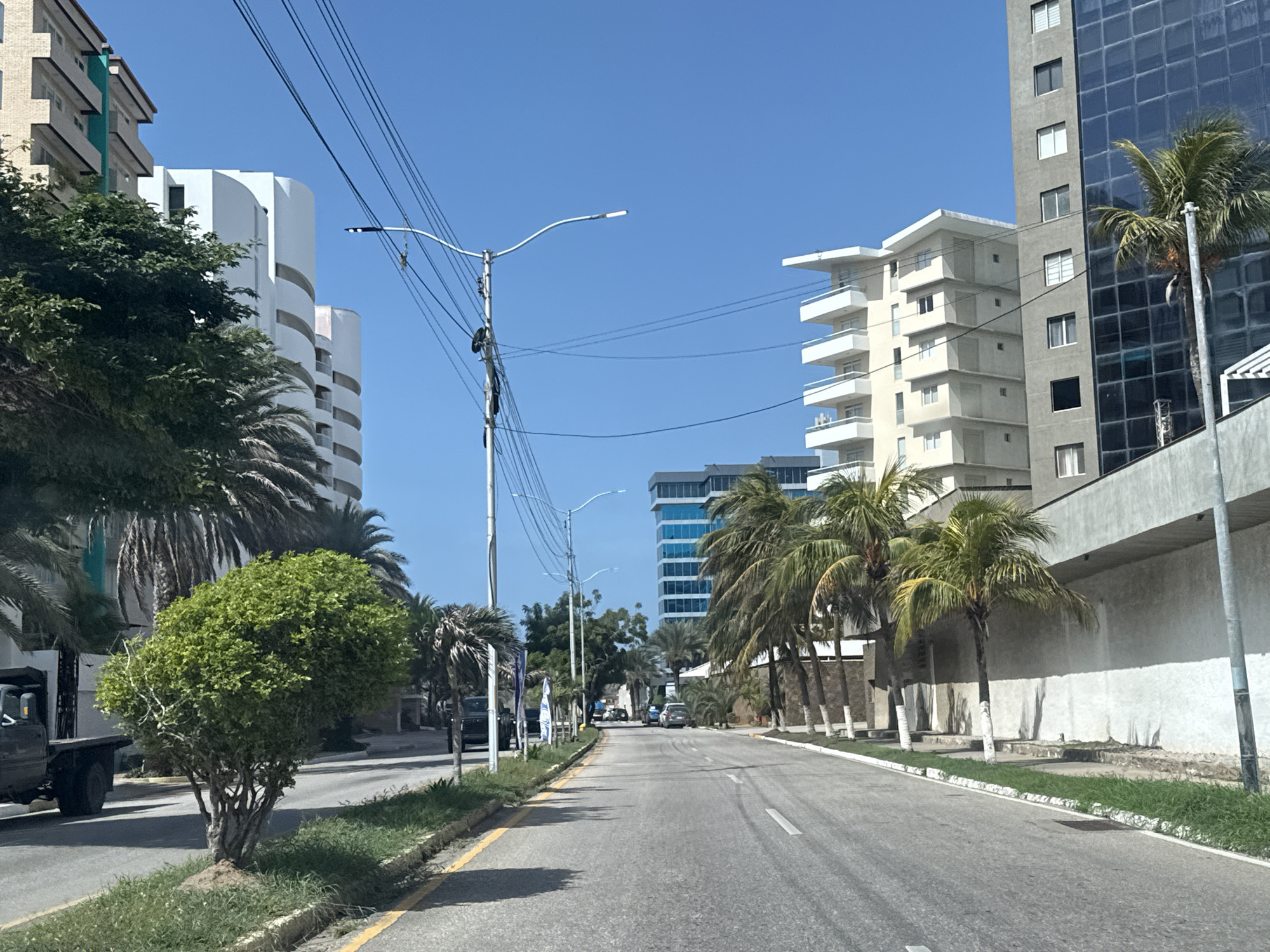 The media file captures a street scene in Pampatar, Venezuela, on a bright, sunny day, indicated by the clear blue sky and strong shadows. The scene depicts a road lined with modern buildings and lush palm trees, suggesting a tropical or subtropical coastal environment.
The primary subjects are the architectural elements: a tall, grey building with a glass facade dominates the right side of the image, while a lighter-colored, multi-story building with white balconies is visible in the middle ground. Other buildings, including a white, curved structure and a lower, cream-colored building with balconies, are also present. Streetlights and utility poles with numerous wires are scattered along the roadside.
Several palm trees, characteristic of the location, are strategically placed along the street, some with their trunks painted white at the base. A small, densely leaved green tree stands prominently in the foreground, near a truck with a flatbed. Other vehicles, including cars and what appears to be another truck, are seen further down the road, suggesting the area is active with traffic.
There is no discernible activity or event taking place, with the scene appearing to be a static view of a street. The overall emotion conveyed is one of a pleasant, bright, and possibly warm day in a well-developed urban or resort area. The presence of cars and buildings indicates a populated and maintained environment. No visible text is present in the image.