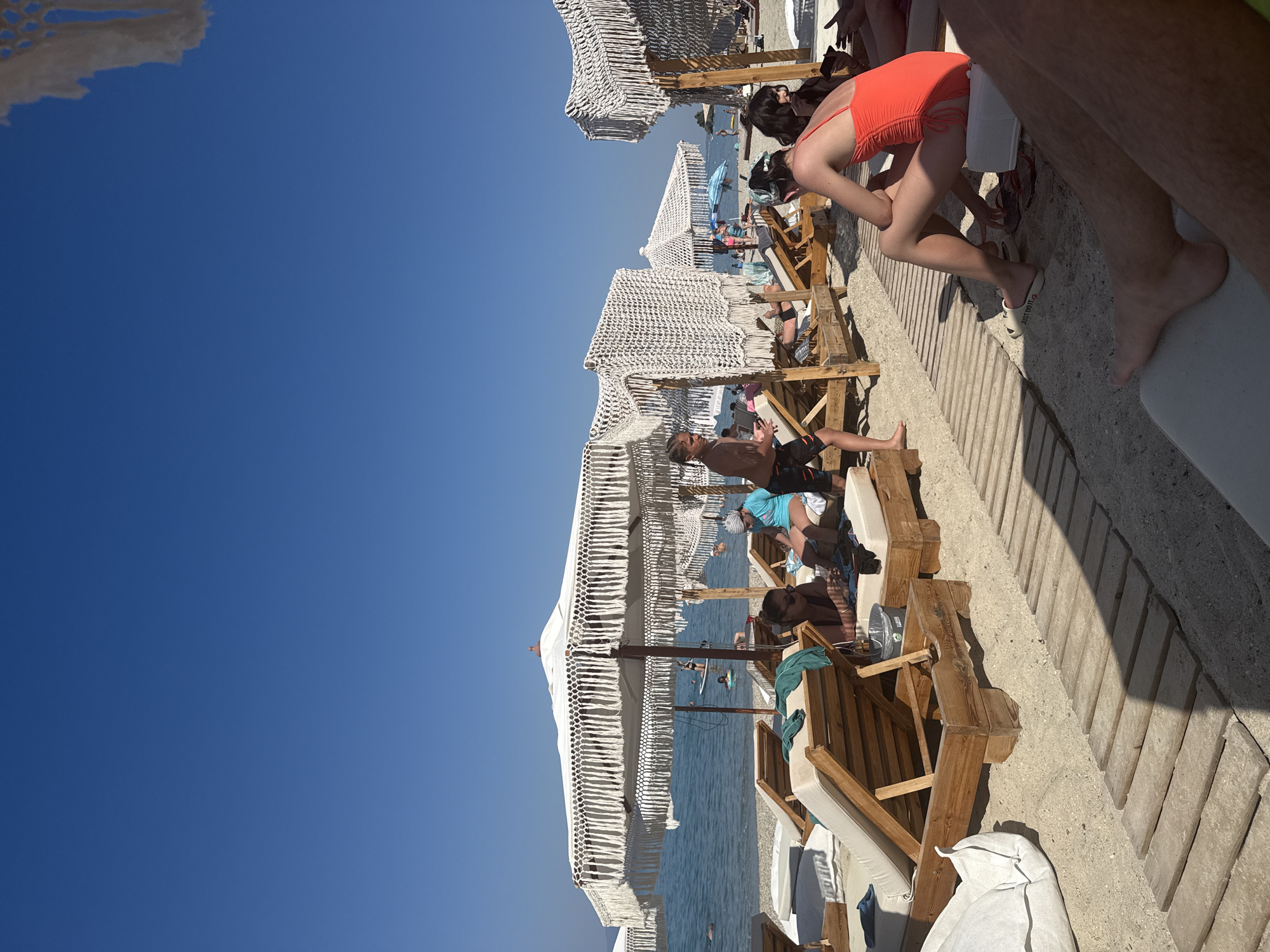The image shows a beach scene in Pallini Municipal Unit, Greece, on a clear, sunny day. The foreground is dominated by sand with a wooden walkway. Several wooden lounge chairs with cushions are visible, some occupied by people. In the mid-ground, there are white umbrellas with decorative fringe, providing shade over lounge chairs. People are seen relaxing on the chairs and walking on the beach. The sea is calm and blue in the background, with a few individuals in the water, some on paddleboards. The overall atmosphere is one of leisure and vacation.