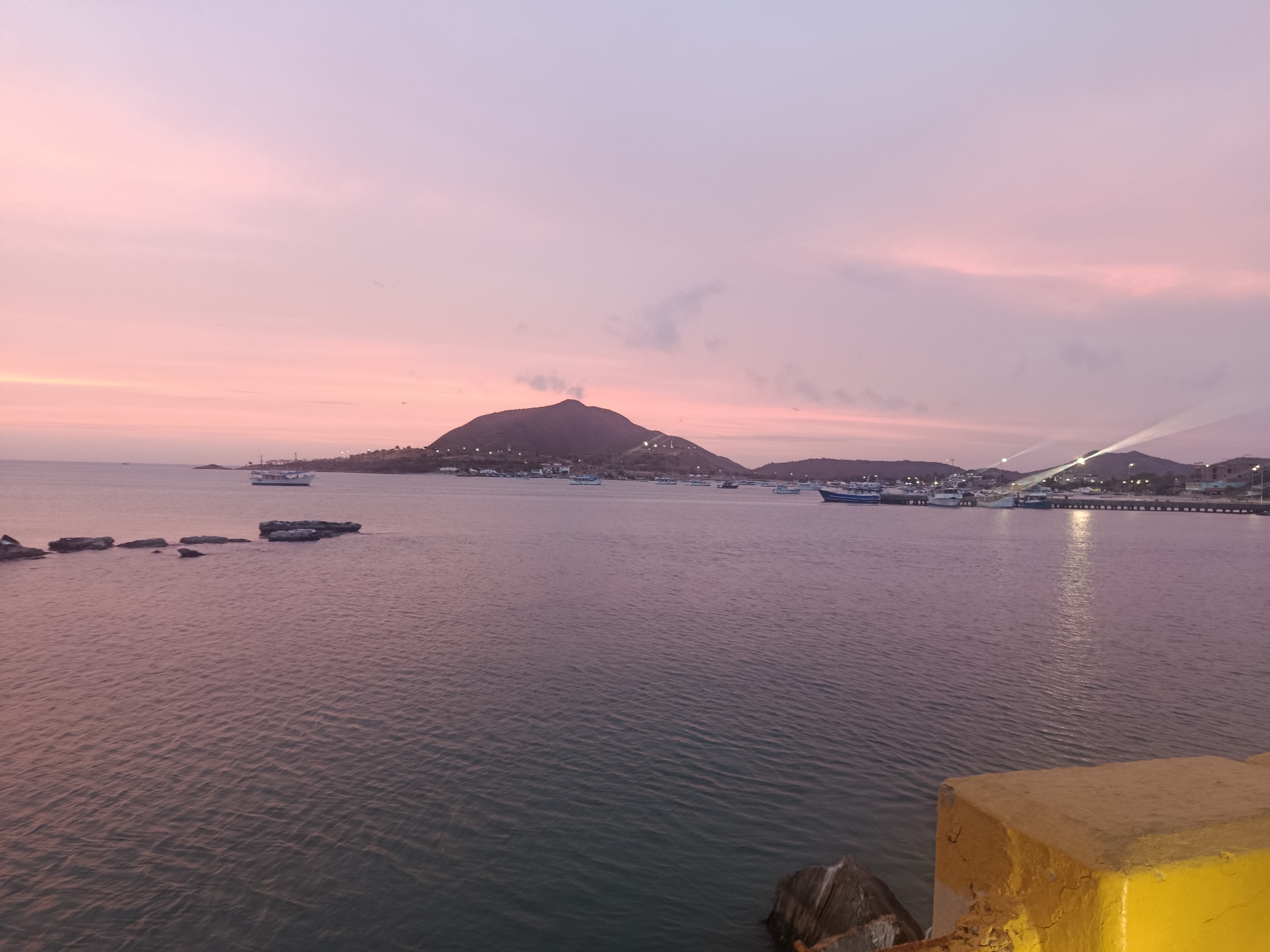 The image shows a view of the bay in Juan Griego, Venezuela at dusk or dawn. The sky is a gradient of pink and light purple. In the center is a large, dark mountain silhouette with lights along its base. The bay is filled with calm water. There are several boats, and a lit pier on the right side of the image. In the foreground, there are dark rocks in the water, and a yellow structure in the lower right corner.