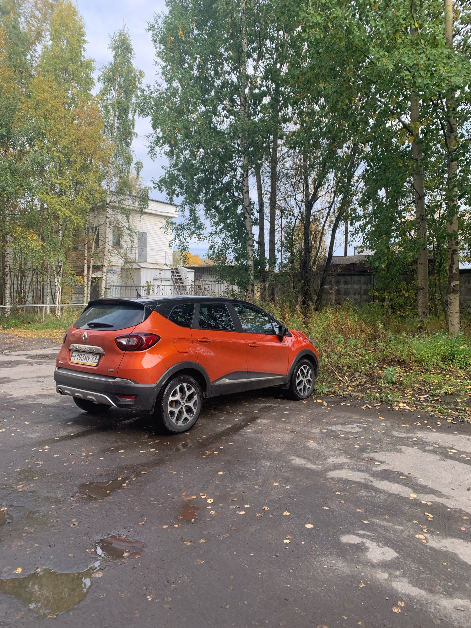 An orange Renault Captur SUV is parked on a wet, asphalt surface littered with fallen yellow leaves. The car is angled towards the right of the frame, with its rear to the viewer. A white building with a light-colored roof and an external staircase is visible behind the car, surrounded by a cluster of trees with yellow and green foliage. To the right of the building and behind the car, a gray concrete wall runs horizontally across the background, with overgrown vegetation in front of it. The sky above is overcast. The scene suggests an outdoor location, likely in Arkhangelsk, Russia, during autumn.