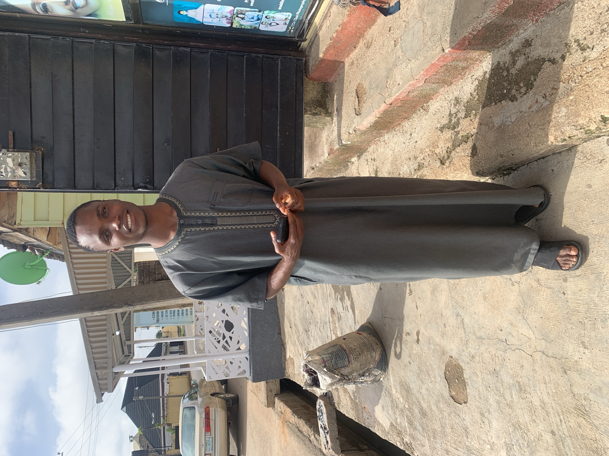 A man stands on a concrete sidewalk in front of a dark metal door. He is wearing a grey tunic and dark sandals. The man is smiling and looking at the camera. Behind him, there is a sign that advertises computer training, online services, and photography. The scene appears to be outdoors during the day, with sunlight casting shadows on the ground. The background shows buildings and a portion of a car. A green satellite dish is visible hanging from a pole to the left of the man. A damaged bag of what appears to be cement is lying on the ground in the foreground. The overall impression is of a casual outdoor scene in an urban environment.