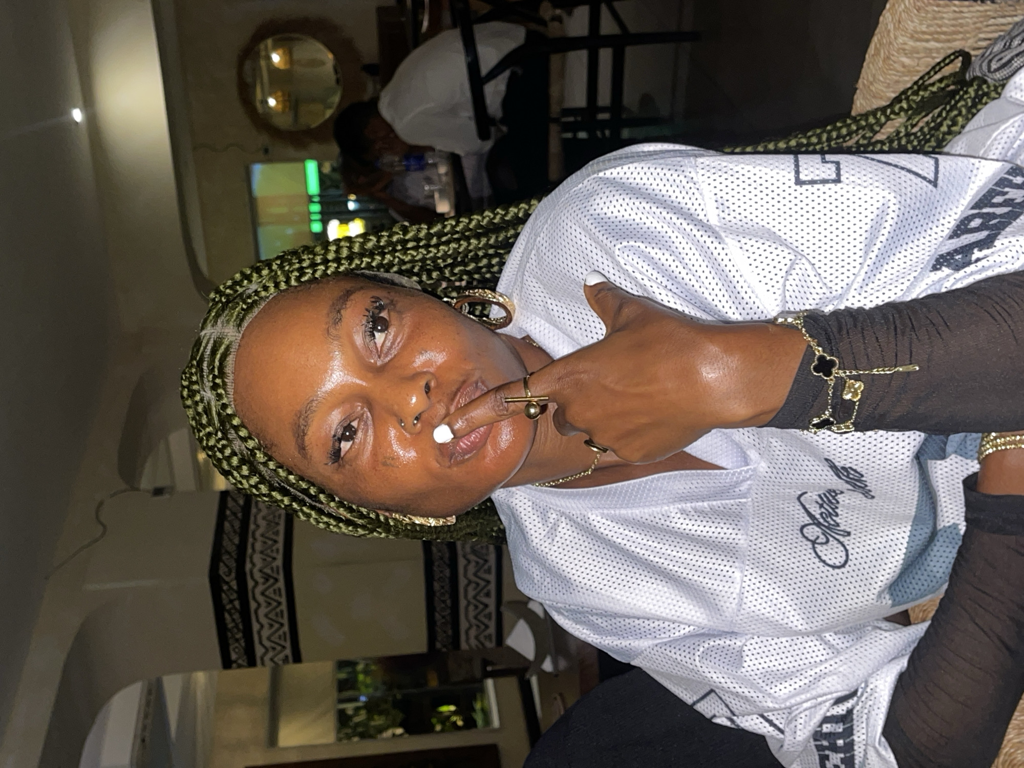 A dimly lit indoor shot captures a Black woman in Ikoyi, Nigeria, wearing a white mesh jersey over a black long-sleeve shirt. She has green braids, gold jewelry, and a stud in her nose. Her finger touches her lips, and she appears to be posing for the camera. In the background, other people are present at tables. A round mirror hangs on the wall, and a fish tank is visible. The environment seems to be a restaurant or bar setting.