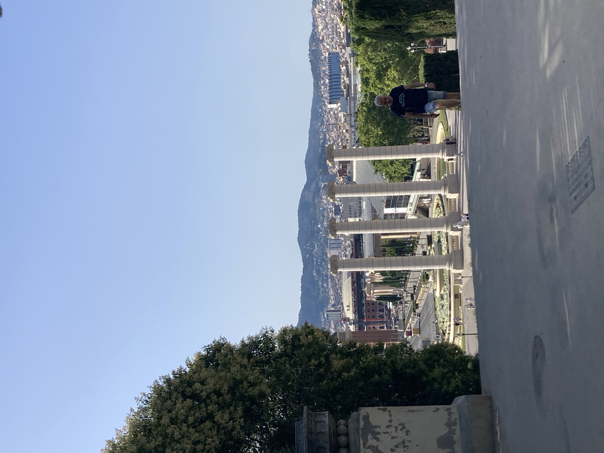 An exterior, eye-level, full shot shows a man walking down a set of steps in a public park in Barcelona, Spain, on a clear day. The man, who is wearing a dark t-shirt with a graphic of a car and blue denim shorts, walks away from the camera toward a fountain and the city skyline. In the background, four large white pillars stand in a row, and beyond them, the city of Barcelona stretches out toward a mountain range. The trees are green and full, and the sky is a clear, light blue. The foreground shows a concrete path, and to the left, a large, partially weathered stone pedestal.