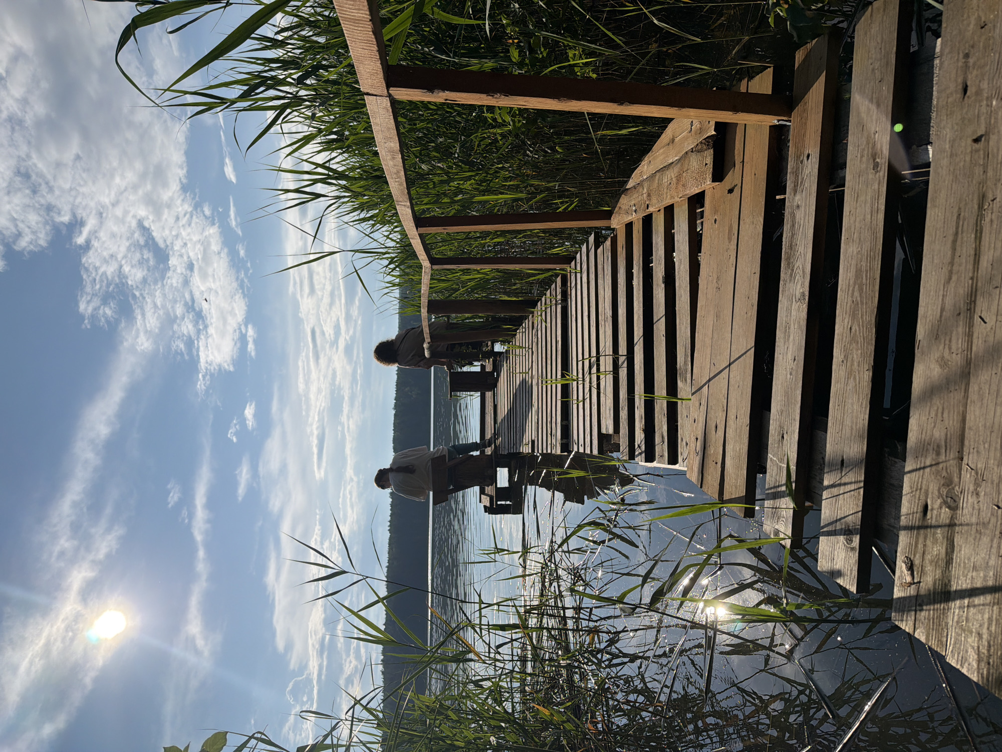 Two individuals, seen from behind, sit on a weathered wooden pier extending into a calm body of water. The pier is constructed of individual planks, some showing signs of age and wear. Tall reeds and grasses grow abundantly along the edges of the water and around the pier. The sun shines brightly in a blue sky dotted with white clouds, casting reflections on the water's surface and creating shadows on the pier. In the background, a distant tree line marks the far shore of the lake. The scene appears to be a natural setting, likely a lake or riverbank, and is located in Votkinsk, Russia.