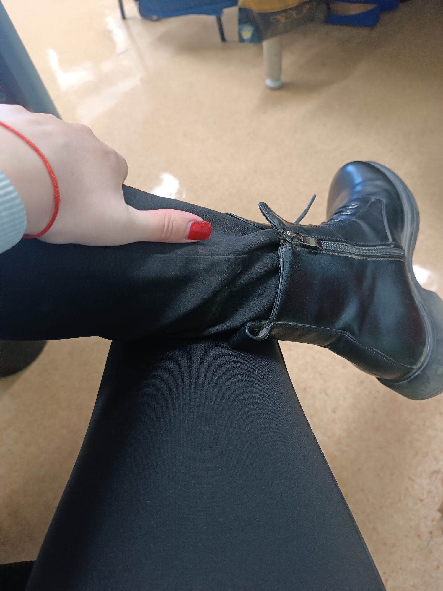 A close-up shot shows a person sitting with their legs crossed, wearing black leggings and a black boot. The person's hand rests on their thigh. The thumbnail is painted red, and a red bracelet is visible on the wrist. The location is Chaikovsky, Russia. The environment appears to be an indoor setting.