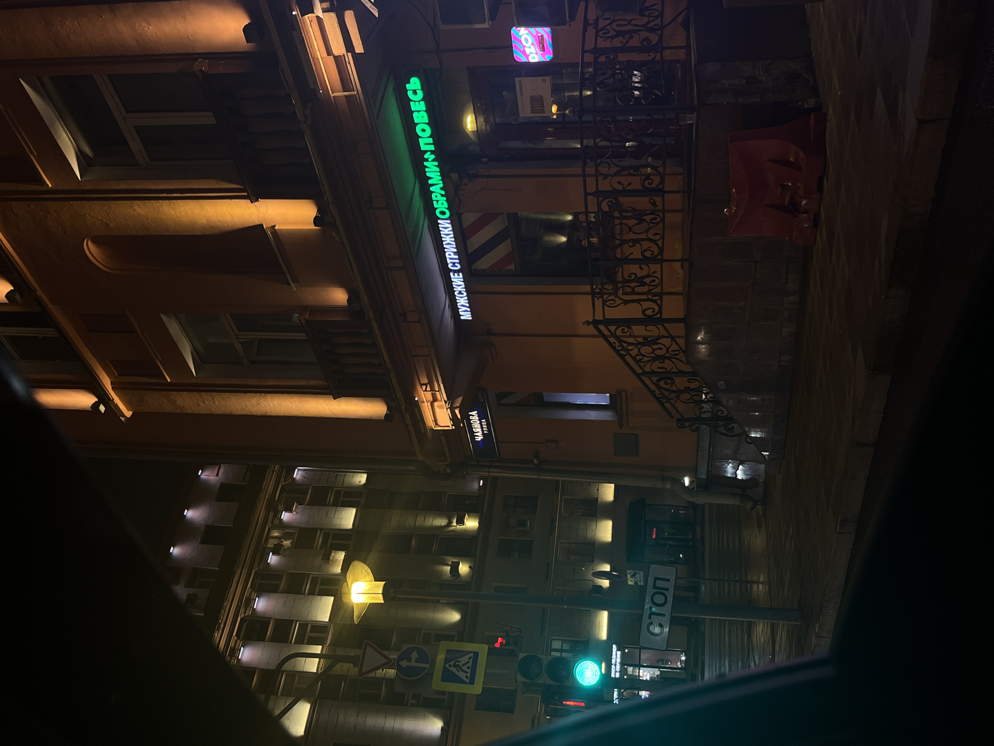 Here's a description of the image:
**General Description:** This is a nighttime shot from a vehicle, likely a car, capturing a street scene in what appears to be a densely built urban environment. The image is taken from an angled perspective, looking upwards at buildings and street infrastructure.
**Location:** The location is Moscow, Russia. This is inferred from Cyrillic text on signs and the architectural style of the buildings, which is typical for the historical center of Moscow. More specifically, the street signs reading "ЧАЯНОВА УЛИЦА" indicate the street is Chayanova Street.
**Details:**
* **Buildings:** Multi-story buildings with lit windows line the street. Some buildings have ornate architectural features.
* **Signs:** Several signs are visible. A green neon sign displays “МУЖСКИЕ СТРИЖКИ ОБРАМИ ПОВЕСЬ” (Men's Haircuts Obrami Poves), advertising a barber shop. There is also an "DZON" advertisement and some signs in the storefronts. Other signs include street signs and a "СТОП" (STOP) sign.
* **Street Infrastructure:** There is a traffic light showing green, indicating that the vehicle has the right of way. There are also several traffic signs.
* **Objects:** The photo is taken through the windshield of a vehicle, part of which is visible at the edges of the frame. Wet pavement suggests it might have recently rained.
* **Environment:** It is dark, with streetlights and building lights providing illumination. The overall atmosphere is urban and active, even at night.
