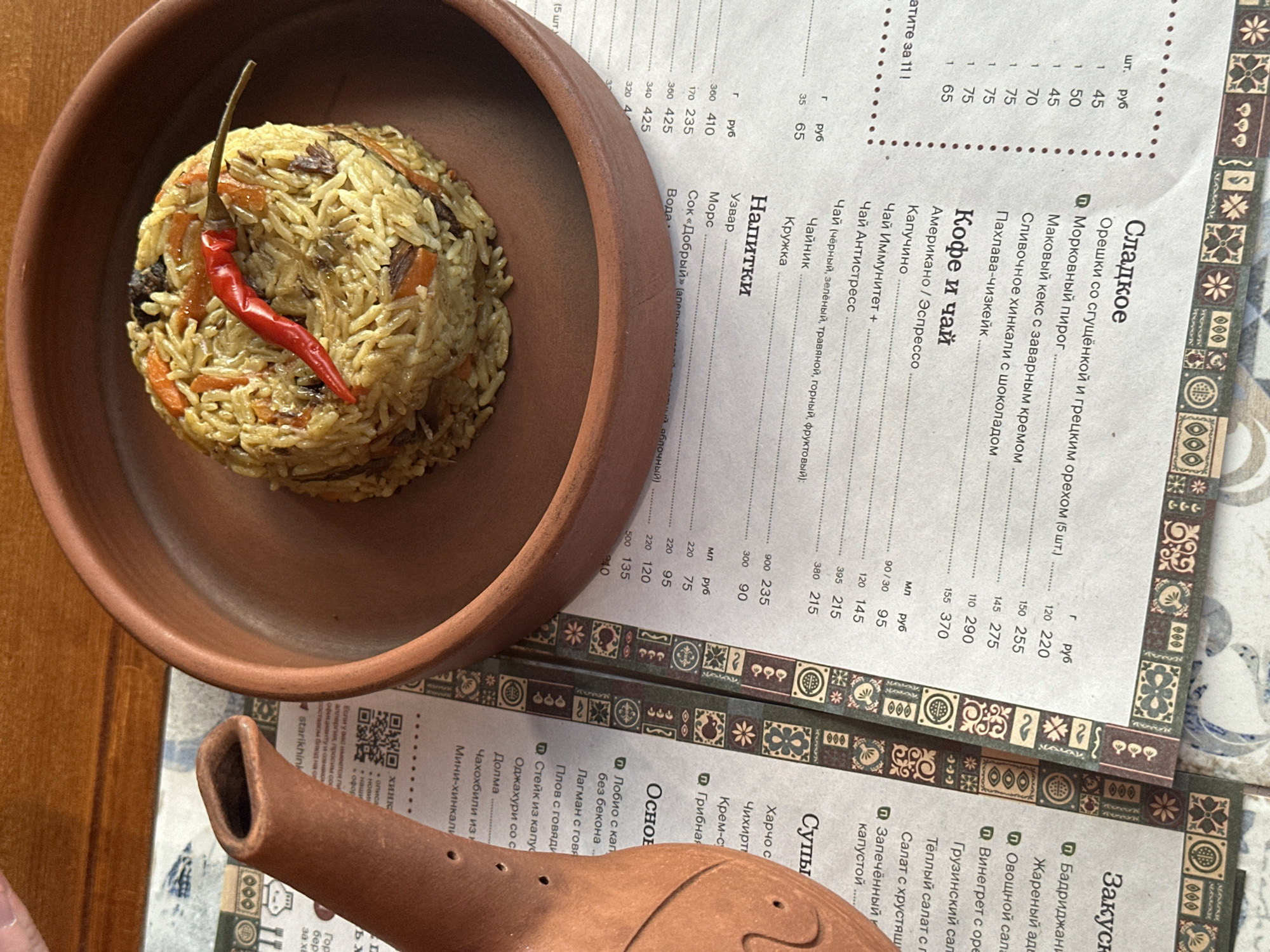 A high-angle shot features a brown ceramic bowl containing a serving of plov, garnished with a red chili pepper, placed on a table alongside a menu. The menu is printed in Russian, including sections for sweets, coffee and tea, drinks, soups, and main courses. A brown ceramic object, possibly part of a teapot or similar vessel, is near the lower left corner of the image. The scene is likely situated in Izhevsk, Russia, given the Cyrillic menu.