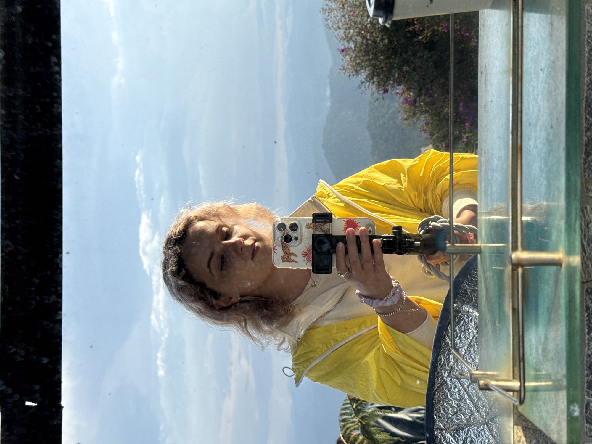 A woman is holding a smartphone with a distinctive decorative case while taking a selfie in front of a mountain vista in Phát Chi, Vietnam. She is wearing a yellow jacket and has blondish hair. The photograph is taken through a transparent barrier or glass panel, which shows the reflection of a telescope and foliage behind the subject. The scenery in the background consists of a cloudy sky and layered mountains. There are metal support structures visible alongside the glass.