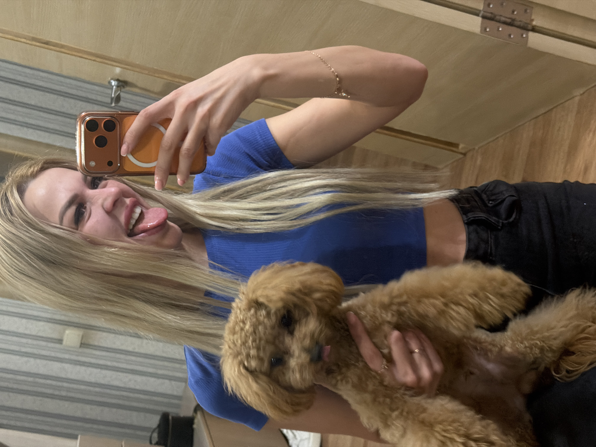 A young woman with blonde hair, wearing a blue cropped top and black jeans, is taking a selfie in a room. She is holding a small, fluffy brown poodle puppy in her left arm, and her tongue is sticking out towards the camera. The puppy is also sticking its tongue out. The woman is holding a phone in her right hand, and a reflection of her is visible in a mirror. The room appears to be a domestic setting, possibly an apartment, with light-colored walls and wooden doors. There is no indication of the city of Kyiv, Ukraine, or any specific location.