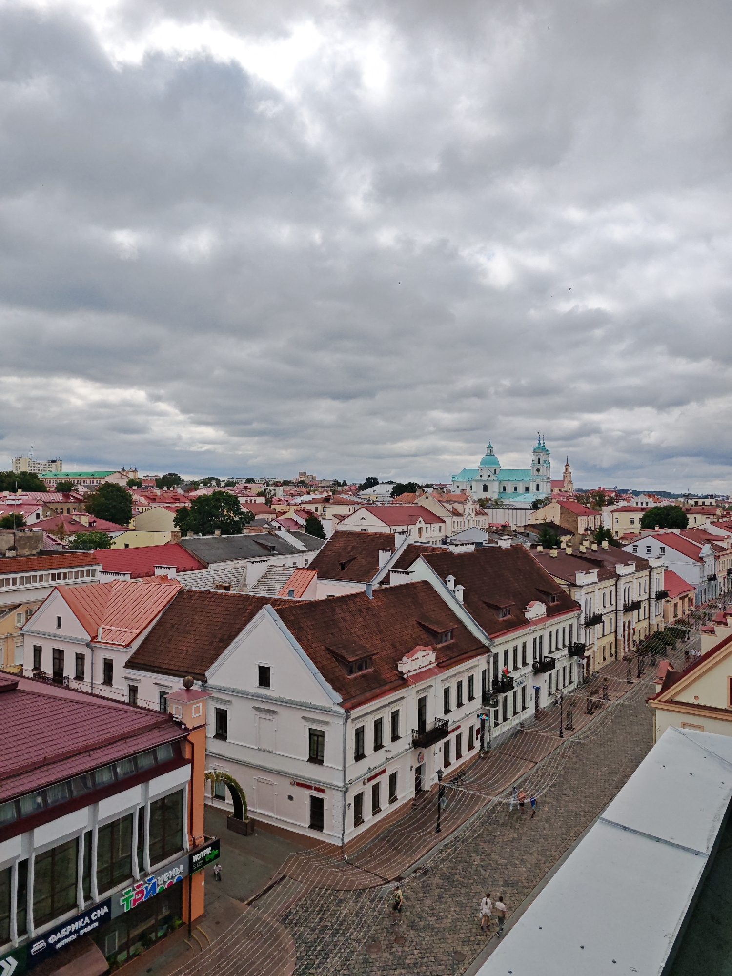 This is an aerial, high-angle, eye-level, medium shot of the city of Hrodna, Belarus, with a focus on the Old Town. The sky is overcast with thick, grey clouds. The foreground is dominated by the rooftops of historic buildings. In the middle ground, a cobblestone street is lined with multi-story buildings with red tile roofs and white facades. There are a few people walking on the street. In the background, a large, pale blue and white church with multiple domes is visible, and beyond that, more buildings and trees complete the cityscape. The image is taken from a high vantage point, likely a building or observation deck, offering a panoramic view of the city.