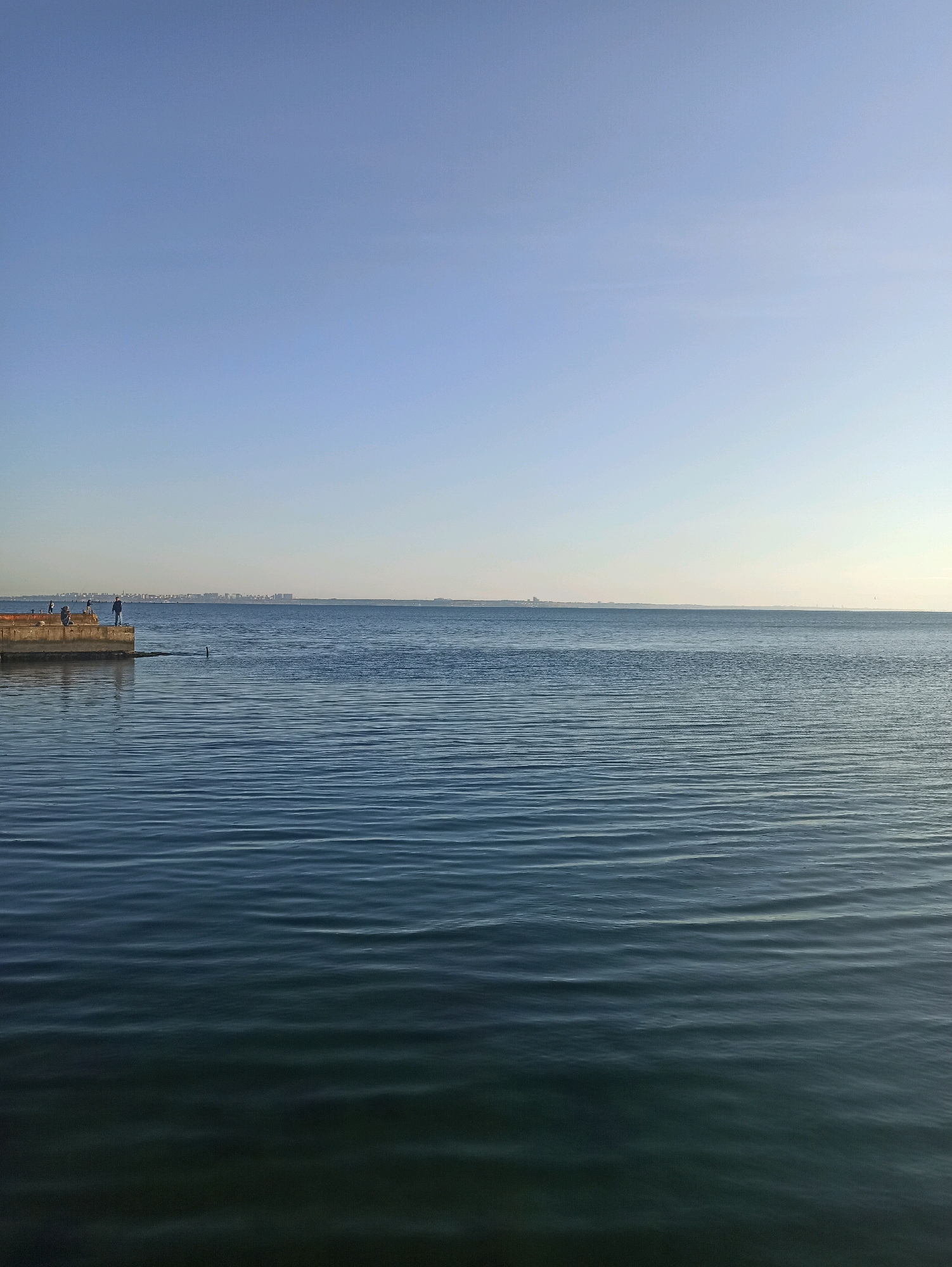 A wide shot captures a tranquil scene on the Black Sea in Odesa, Ukraine, under a clear, pale blue sky. In the foreground, the deep blue water exhibits gentle, rhythmic ripples that create a textured, undulating surface. On the left side, a concrete pier juts out into the water. On the pier, three individuals are visible: two are seated or crouching, and one person stands, appearing to be fishing. The figures are small in the frame, suggesting they are at a considerable distance. Beyond the pier, a distant cityscape of Odesa stretches across the horizon, marked by low-rise buildings. The water extends to meet the horizon, creating a distinct separation between the sea and the sky. A dark, elongated object, possibly a buoy or a small craft, is partially submerged in the water to the right of the pier, creating a subtle interruption in the water's surface. The overall atmosphere is one of quietude and calm.