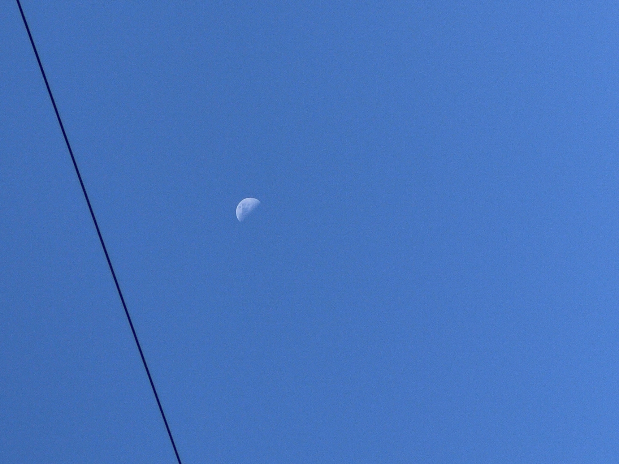 The image captures a clear, vibrant blue daytime sky. In the upper central area of the frame, a distinct half-moon is visible, showcasing some surface texture and shadow details. A dark, thin line, appearing to be a power cable or utility wire, cuts diagonally across the left side of the photograph, extending from the top-left corner downwards towards the bottom-center. The sky is entirely free of clouds, indicating clear weather conditions. No specific geographical landmarks, people, or objects other than the moon and the wire are present. The scene depicts a quiet, open sky during daylight hours, with the moon appearing as a celestial object in the clear atmosphere above Chaikovsky, Russia. No visible text is present in the image.