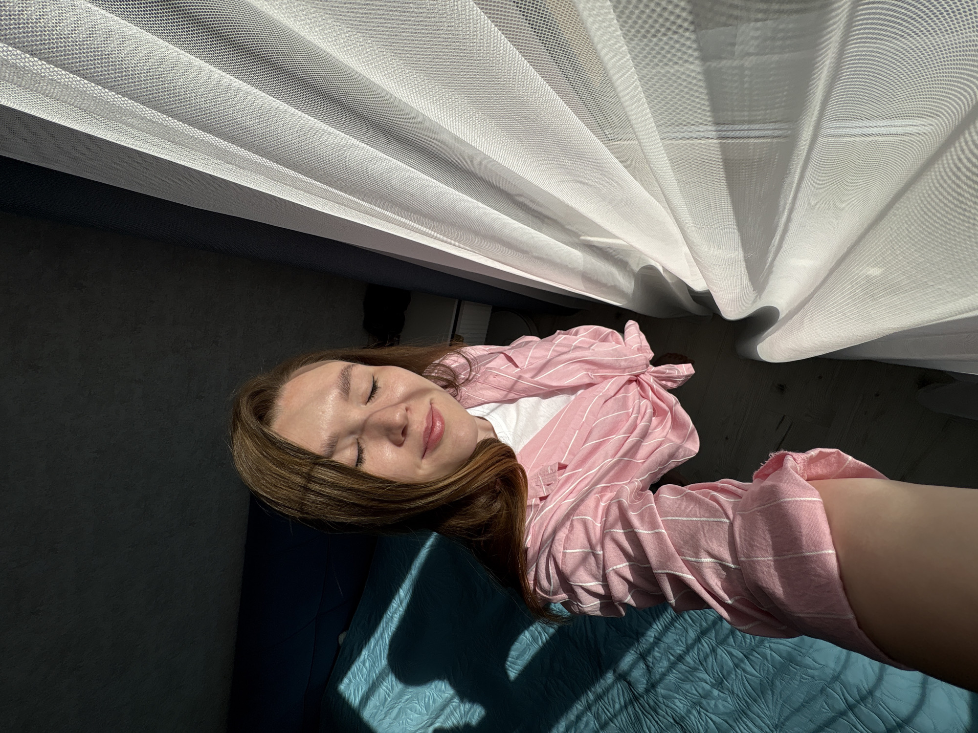 A young woman lies on a blue quilted bedspread in a room bathed in light. She wears a white t-shirt and a pink and white striped long-sleeved shirt. Her eyes are closed, and she appears relaxed. A white curtain hangs in the background, filtering the sunlight. The room's walls are a dark gray color. The location is unspecified, and there is no evidence to suggest that this image was taken in Izhevsk, Russia.