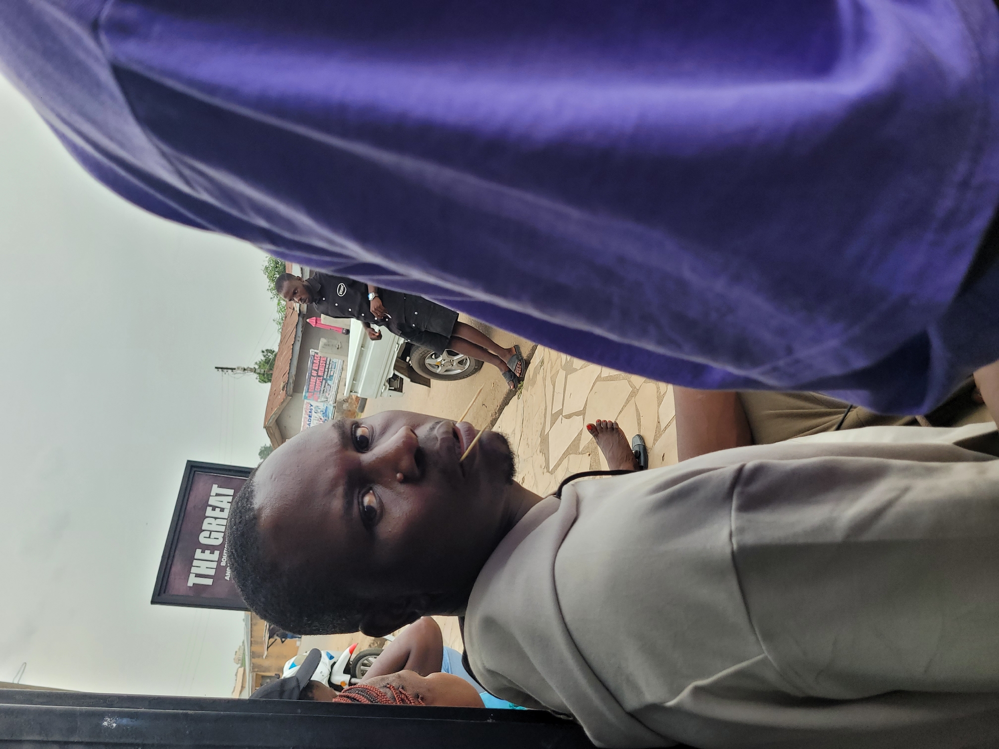 A low-angle outdoor shot taken during the day in what appears to be Jos, Nigeria, features a man as the primary subject. He's looking directly at the camera with a toothpick or small twig in his mouth. He is wearing a light-colored shirt and is partially obscured by what seems to be a purple garment, possibly clothing, draped above.
In the background, visible between and around the figures, is a street scene with buildings and vehicles. A sign reading "THE GREAT" is partially visible to the left. A second person, dressed in black with an "INTEL" logo, stands near a white car. A third person is partially seen lying down. The sky is overcast, suggesting possible rain. The flooring consists of paving stones.