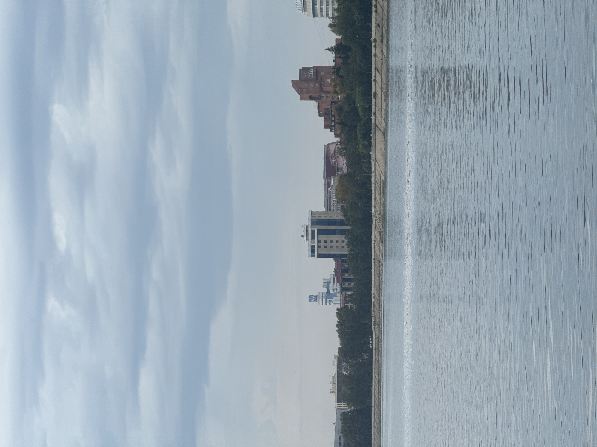 The image shows a panoramic view of a city skyline across a body of water. The sky is overcast with light gray clouds. The water in the foreground is calm with visible ripples. In the background, a tree line separates the water from the city. Various buildings of different architectural styles and heights are visible, including modern high-rises and older brick structures. Some of the buildings have visible windows and architectural details. The general impression is that of a city situated on a waterfront. The scene appears to be captured from a distance, likely across a river or lake. This view is characteristic of the Irkutsk Reservoir (also known as the Irkutsk Sea) in Irkutsk, Russia, looking towards the city's developed waterfront. No people are visible in the image.