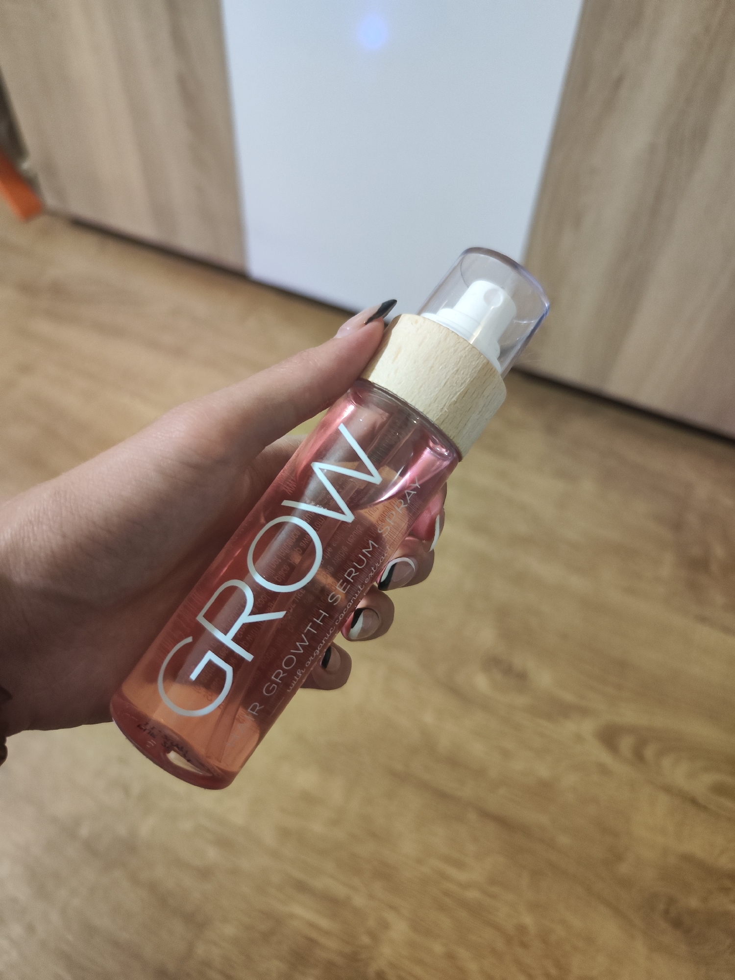 A person's hand, with black and white nail art, holds a clear, pink-tinted spray bottle with a wooden collar and a clear plastic cap. The bottle has white lettering that reads "GROW HAIR GROWTH SERUM SPRAY" and smaller text below that appears to say "with organic coconut extr". The background is a wooden floor and a blurred section of a wardrobe. The scene appears to be indoors, possibly in a residential setting. There is no indication of the city of Sofia, Bulgaria.