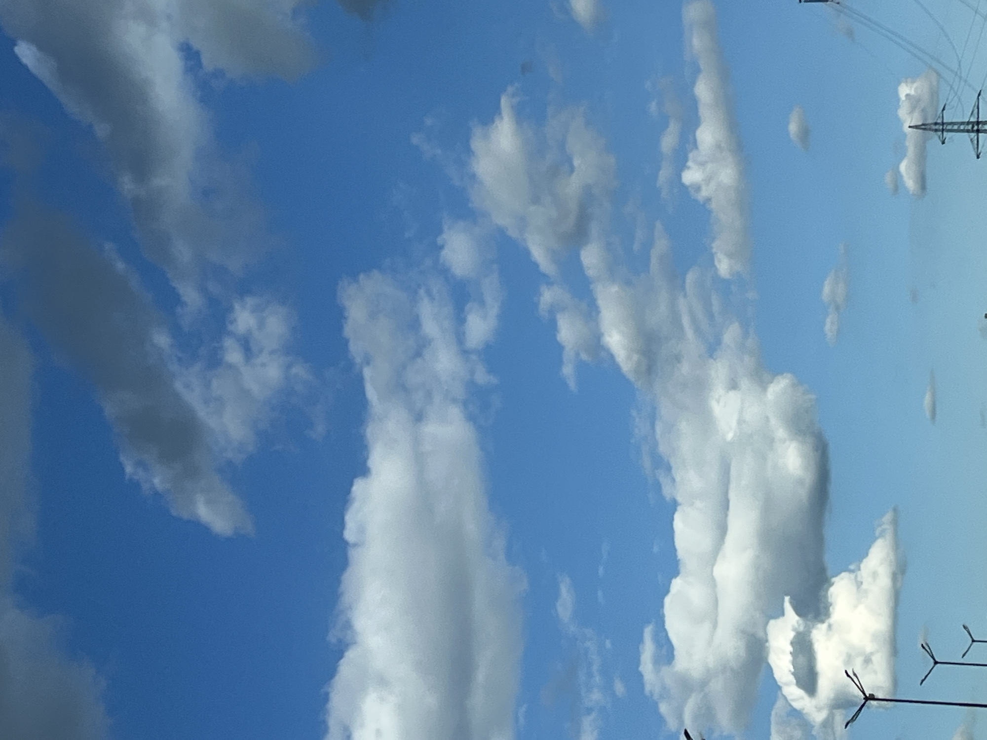 This image captures a dynamic sky filled with a variety of clouds over an unspecified urban landscape, potentially in the city of Unknown, Unknown. The dominant feature is the brilliant blue sky, partially obscured by a mix of cumulus and darker, more ominous clouds. These clouds are scattered across the frame, some appearing fluffy and white, while others are tinged with gray, suggesting depth and movement.
In the lower portion of the image, the silhouettes of man-made structures are visible, hinting at the urban environment below. These include several tall lampposts with distinctive, multi-armed fixtures, typical of street lighting. Further to the right, a power line pylon with a lattice structure is discernible, along with the faint outlines of power lines stretching across the sky. The overall impression is one of an overcast but bright day, with the vastness of the sky and the subtle presence of human infrastructure dominating the scene. There are no people or animals visible in the image.