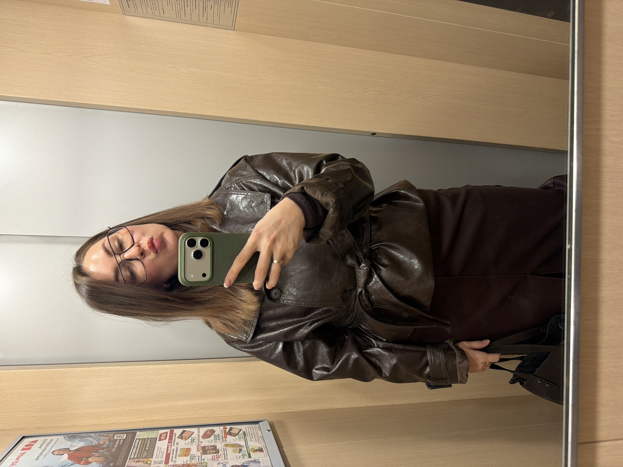 The image shows a woman taking a selfie in a mirror. She is wearing glasses, a brown leather jacket, and a brown dress. She is holding a green smartphone. A purse is visible in the reflection of the mirror. A poster with Russian text is on the bottom left corner of the image. This poster likely indicates the location being in Russia. A poster on the poster has the word "Ижевск," or "Izhevsk," which can be translated into English. It is a city in Russia. The walls are a beige or light wood color.