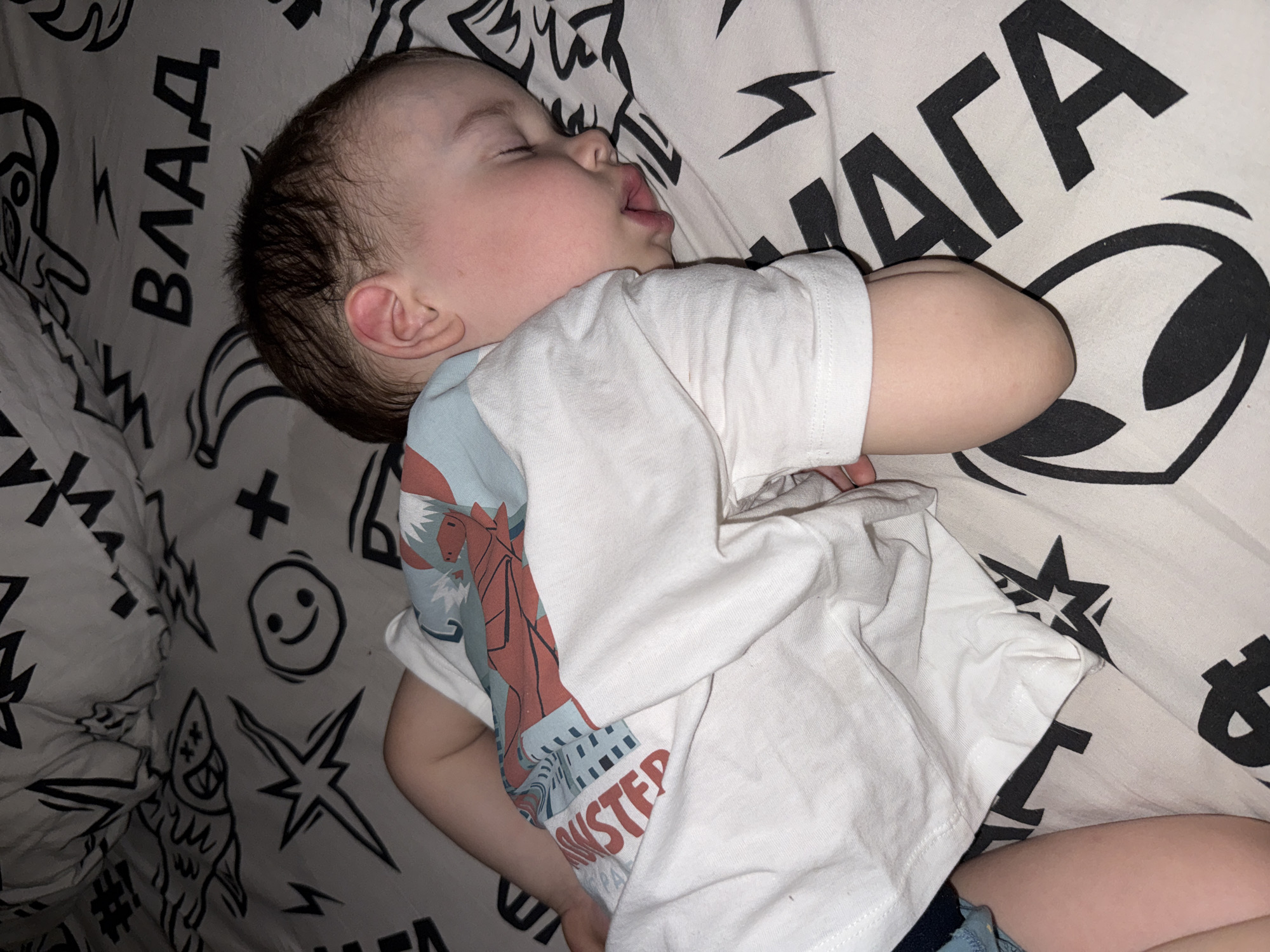 A toddler lies asleep on patterned bedding. The child is lying on their side, with their eyes closed and mouth slightly open. They are wearing a white t-shirt with a graphic design on one side. The bedding has a black and white pattern with various designs including words in Cyrillic script, smiling faces, stars, and an alien head.