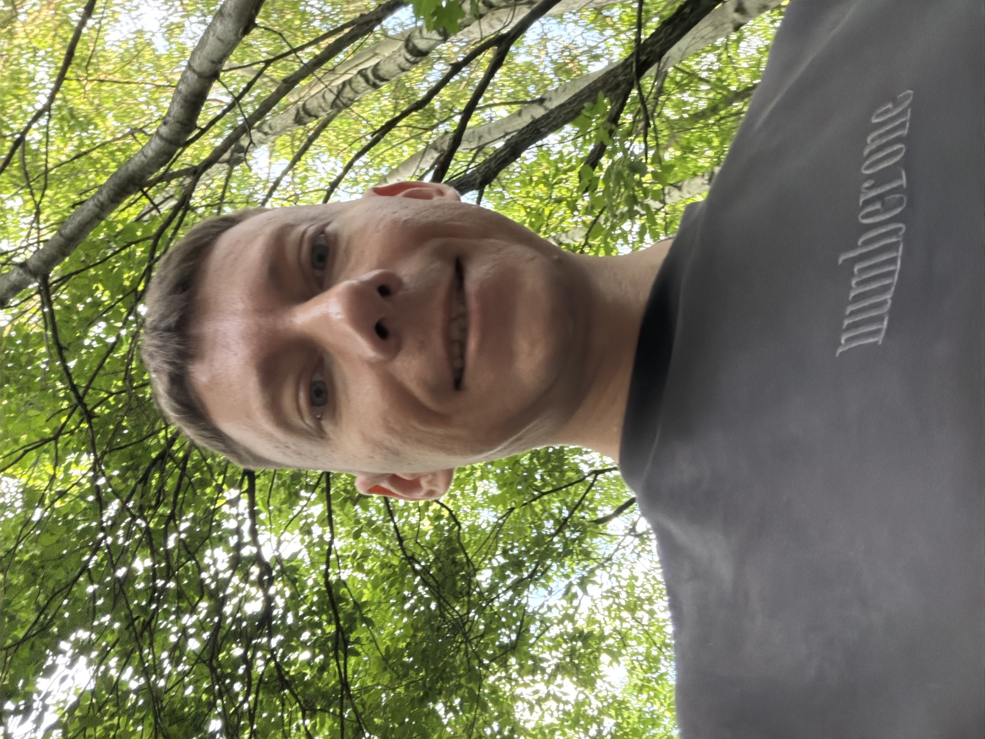 A close-up, selfie-style shot features a man in his early 30s with short, buzzed brown hair, blue eyes, and fair skin. He is smiling at the camera, with his teeth visible, and has light stubble on his face. He is wearing a dark gray t-shirt with the word "number one" embroidered in white on the left side of the chest. The background is filled with lush green foliage and the trunks of birch trees, suggesting an outdoor setting, possibly a park or forest. The lighting indicates it is daytime. No other individuals or objects are visible in the frame. The scene is consistent with a casual outdoor portrait taken in a wooded area within Moscow, Russia.