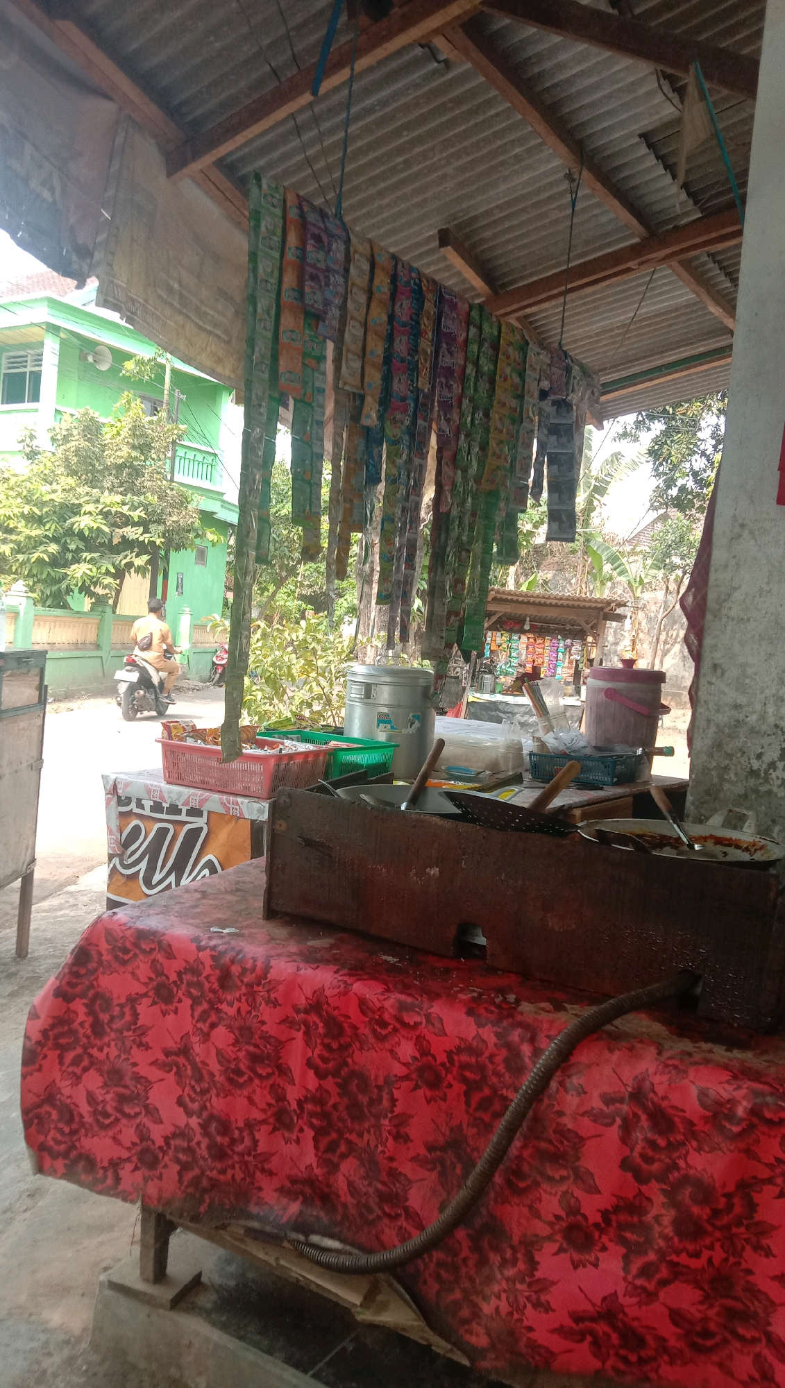 The image depicts an outdoor food stall in Ngempit, Indonesia. The stall is covered by a corrugated metal roof supported by wooden beams. Hanging from the roof are numerous small, colorful packets, likely containing snacks or condiments. In the foreground, a table is covered with a red cloth featuring a black floral pattern. On the table are various cooking utensils and containers, including a large metal pot, a wok, and a blue basket. A person is seen riding a motorcycle in the background, passing by a bright green building. The environment appears to be a typical street scene in a residential area, with lush green trees and other buildings visible. The lighting suggests it is daytime, with the sun casting shadows.