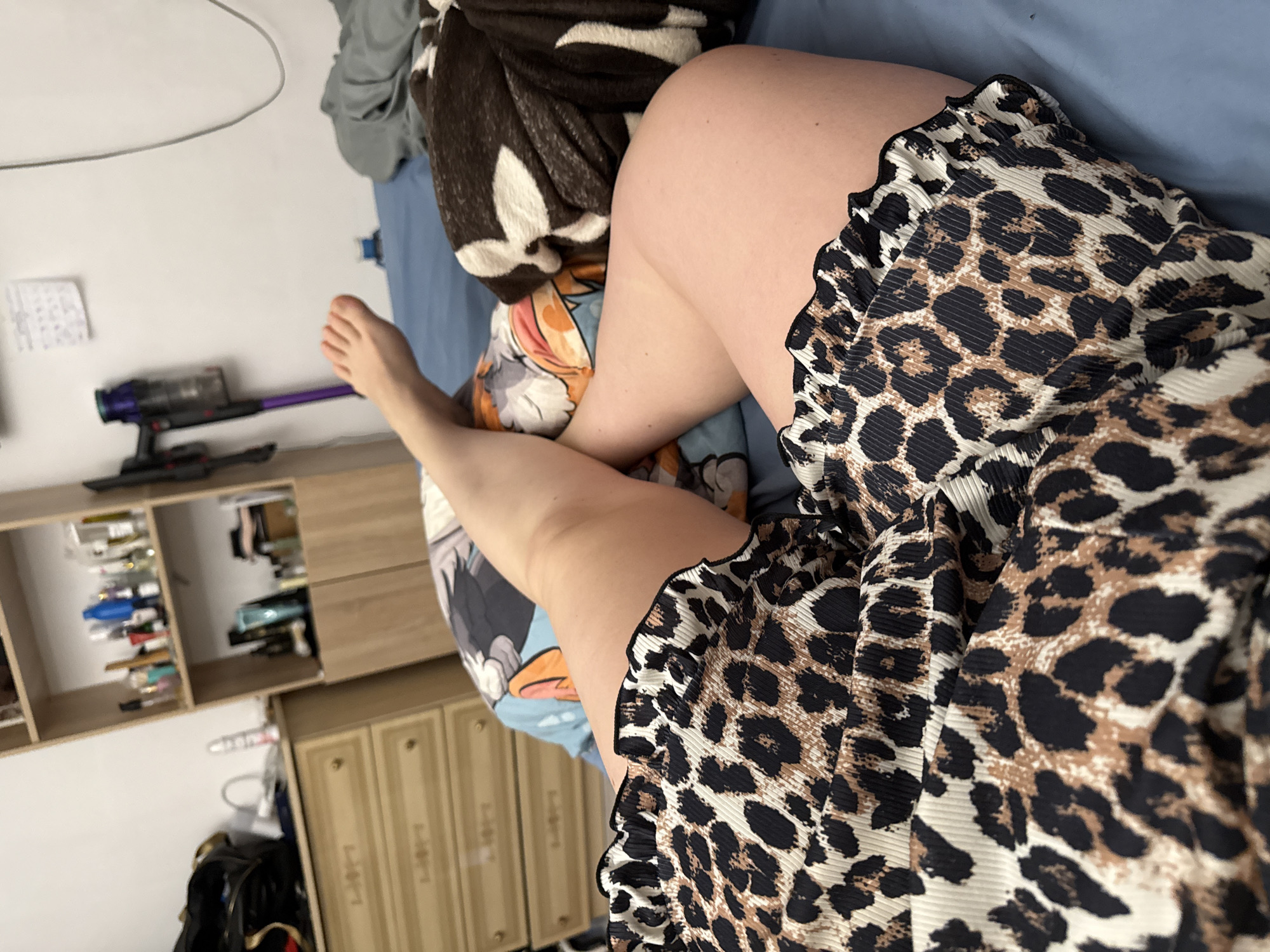 A top-down shot depicts a person sitting on a bed. The individual is wearing an animal print garment. Part of their leg is visible. The bed has blue sheets, with additional blankets and pillows with cartoon designs. A vacuum cleaner is on a shelf. A dresser is adjacent to the shelf. The location is not apparent. The photo was likely taken indoors in a bedroom.