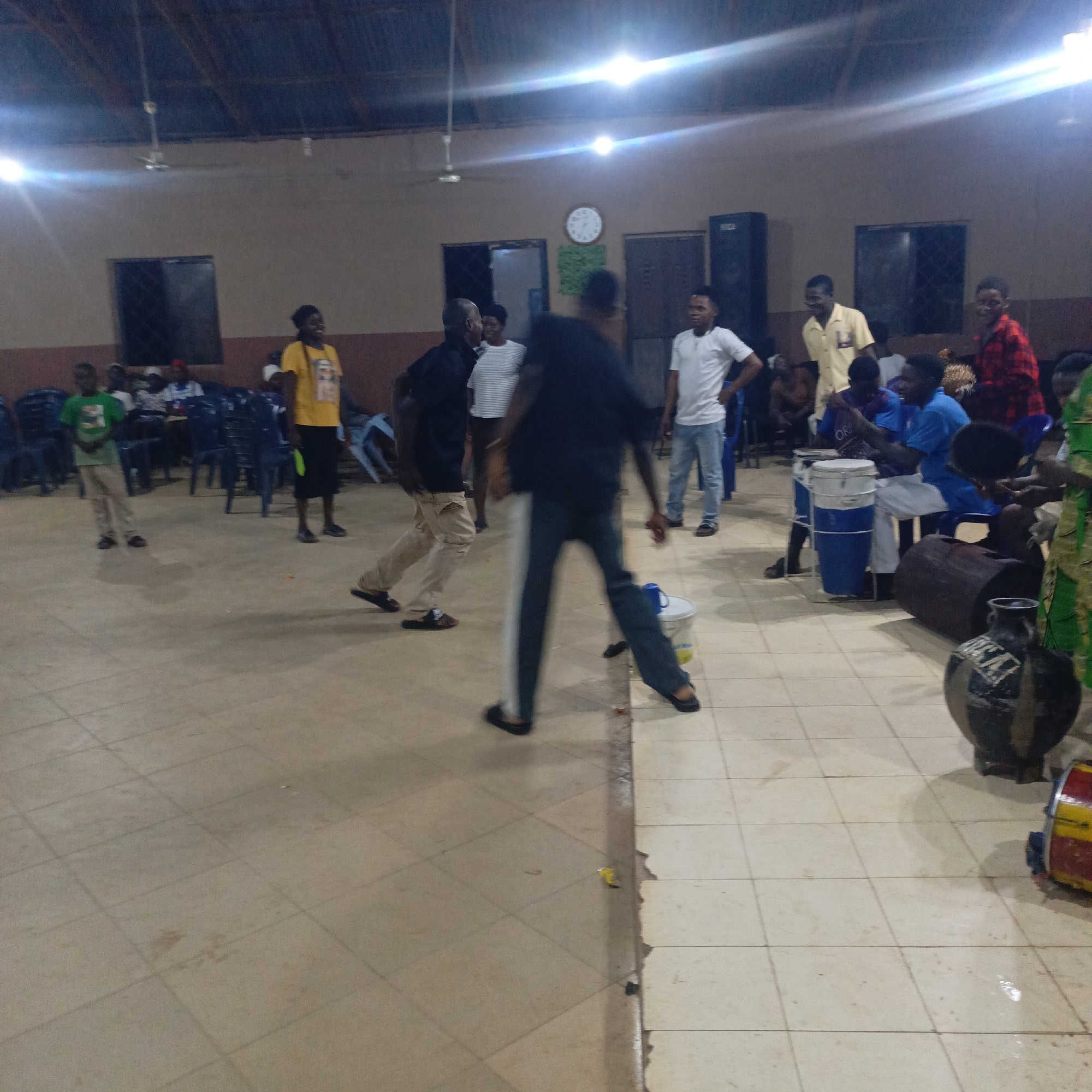 Akwanga, Nigeria. A large room with tile flooring and overhead lighting. A group of people are present. Some are seated in blue plastic chairs, while others stand or dance. Some people are playing drums. There are windows, doors, a clock on the wall, and a speaker.