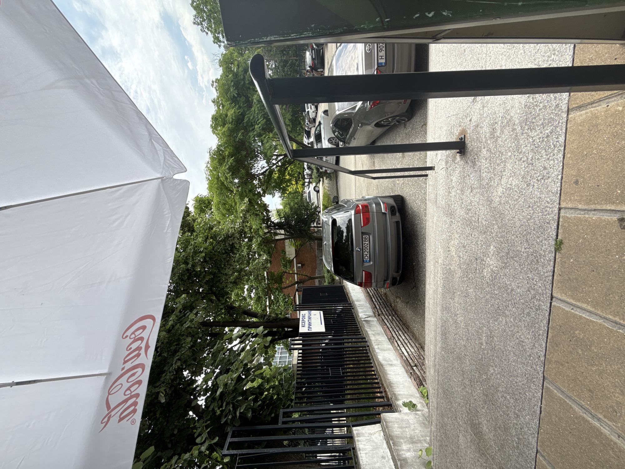 The image depicts an outdoor street scene, likely in an urban area, under a bright, partly cloudy sky during the daytime. In the left foreground, a large white umbrella with the distinctive red "Coca-Cola" logo is prominently featured. To its right, a grey, speckled granite pavement or sidewalk extends, bordered by a black metal railing.
Beyond the railing, a street or parking area is visible, with several cars parked along the curb. A grey SUV, likely a BMW X5, is parked closest to the viewer on the right, displaying a license plate reading "CH 2002 BB". Further down the street, other cars are parked, including a silver sedan with a partially visible license plate starting with "H 907". Lush green trees and foliage line the street behind the parked vehicles, obscuring most of the background buildings, though hints of brick structures can be seen.
Attached to the black metal railing is a white sign with Cyrillic text. The text clearly reads "КОПИРНЕ" (KOPIRNE) and "ПРИНТИРАНЕ" (PRINTIRANE), which translates to "Copying" and "Printing" services. No people are visible in the scene. The presence of the Coca-Cola umbrella suggests a cafe or commercial establishment nearby. The license plates and Cyrillic text are consistent with the provided location context of Plovdiv, Bulgaria.