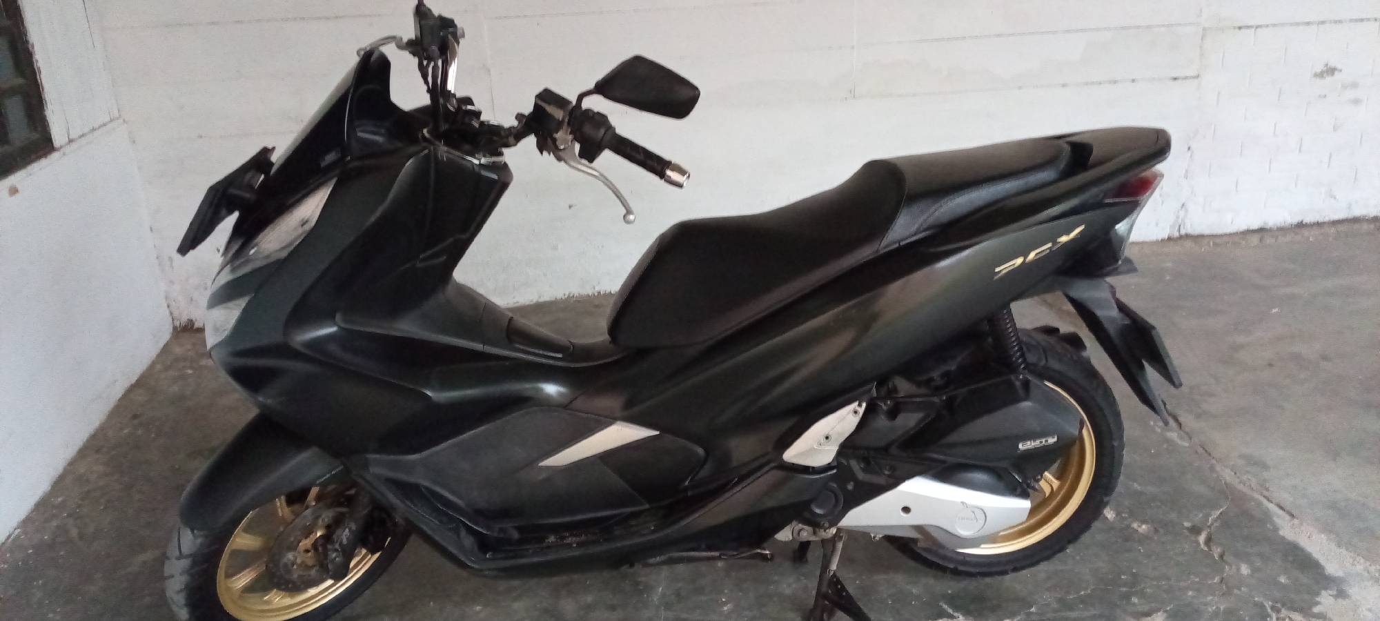 The image shows a black Honda PCX scooter parked indoors. The scooter is angled towards the right, with its front wheel on the left side of the frame and its rear wheel on the right. The scooter has a sleek, modern design with a black body and gold accents on the rims. The seat is black and appears to be made of leather.
The scooter is parked on a concrete floor, and the background consists of a white, slightly textured wall. The lighting suggests it is daytime, but the exact time is difficult to determine due to the indoor setting. There are no people visible in the image.
Notable details include the "PCX" logo in gold lettering on the side of the scooter. The scooter appears to be in good condition, with no visible damage. The setting seems to be a garage or a covered parking area, given the plain walls and concrete floor. The overall impression is one of a well-maintained vehicle in a functional, nondescript environment. The location context of Lhokseumawe, Indonesia, does not provide any specific visual cues within the image itself.