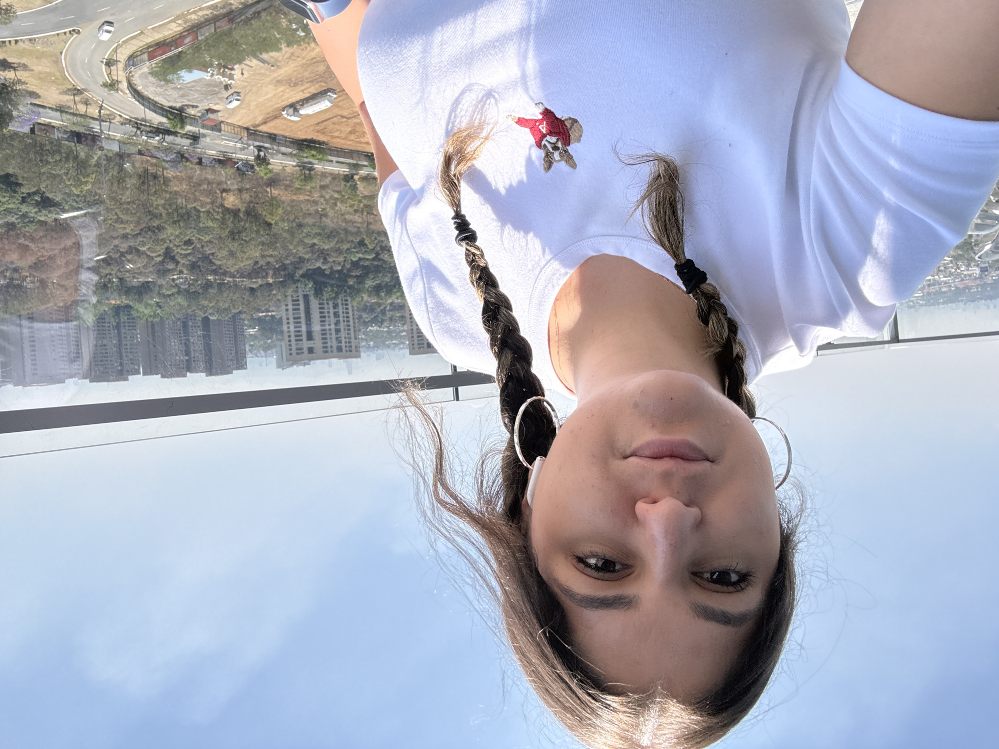 A woman with braided brown hair, fair skin, and wearing large hoop earrings and a white t-shirt with a small image of a red-jacketed figure is facing the camera. She is standing indoors, overlooking a cityscape, including buildings, trees, and roads with vehicles. The image is taken from a high vantage point, possibly a building or structure in Mexico City, Mexico.