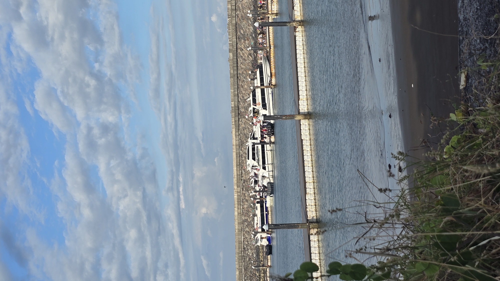 The image depicts a harbor scene likely in Denpasar, Indonesia, based on common features of the area. Several tourist boats are docked along a concrete pier. A row of concrete blocks lines the harbor edge. People are visible on some of the boats and along the pier. The harbor water is relatively calm. A beach borders the harbor to the right of the pier. In the foreground, there is some vegetation along the edge of the harbor. The sky is blue with scattered clouds.