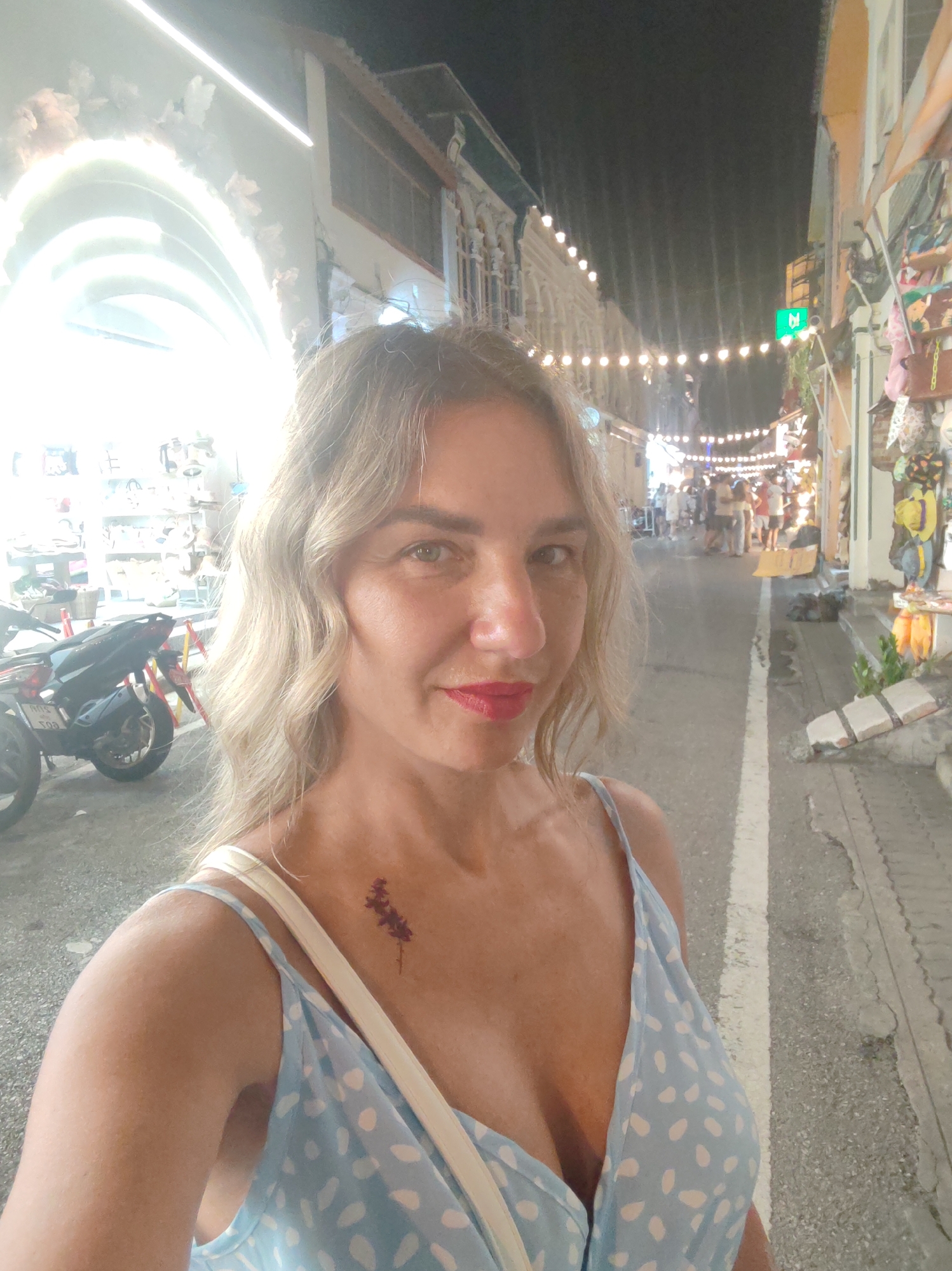 The image is a nighttime selfie of a woman on a bustling street, likely in Phuket City, Thailand. The primary subject is a woman with medium-length wavy blonde hair, light-colored eyes, and red lipstick, looking directly at the camera. She wears a light blue spaghetti-strap dress with white polka dots, and a white strap from a bag is visible over her right shoulder. A small, dark red, floral-like tattoo or sticker is present on her upper chest/collarbone area.
The background depicts a vibrant outdoor scene illuminated by artificial lights. To the woman's left, a bright, overexposed white arched structure glows intensely, and a motorcycle is parked nearby. To her right, the street extends into the distance, lined with buildings that appear to be shops or stalls. String lights are strung across the street overhead, casting a warm glow on the scene. A crowd of people is visible further down the street, suggesting a lively market or pedestrian area. The dark sky confirms it is nighttime. The street is paved and features a white dividing line. A green square sign with a white symbol, possibly an 'M', is visible above the crowd on the right. Faint Thai script is visible on the license plate of the motorcycle behind her. The scene conveys a busy and energetic evening atmosphere.