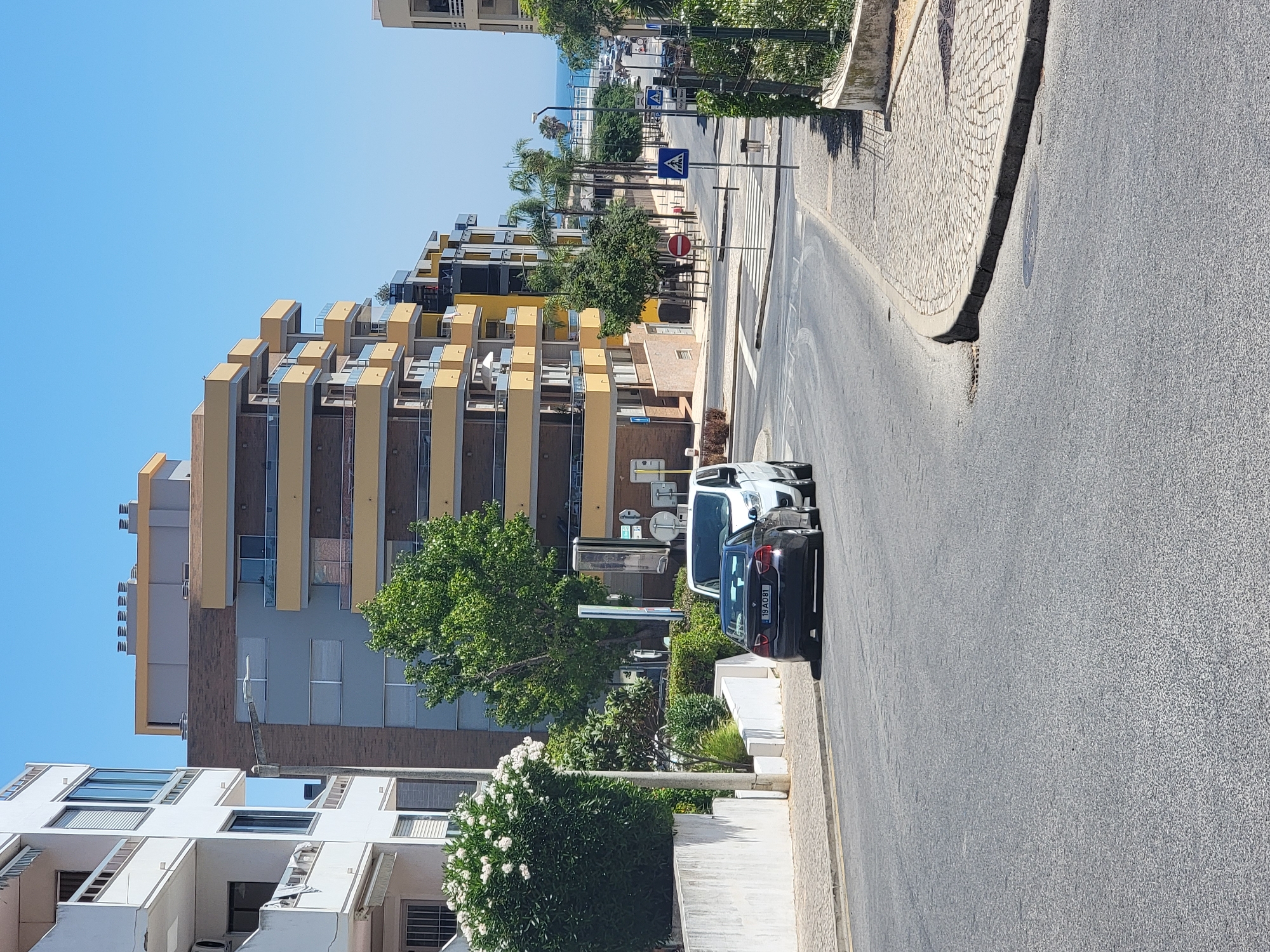 The image shows a street scene in Quarteira, Portugal, on a sunny day. In the foreground, a dark-colored sedan with the license plate "19 AO 81" is parked on the right side of the road, followed by a white van. The road curves to the right, leading towards a pedestrian crossing with a blue and white "X" symbol. Across the street, there are several multi-story buildings with yellow balconies and what appear to be apartments or hotels. Lush green trees and bushes are visible, some of them in full bloom with white flowers. In the background, palm trees and other vegetation line the street, and beyond that, the ocean is visible under a clear blue sky. Several street signs, including pedestrian crossing signals and a red circular "no entry" sign, are placed along the road. The overall impression is a well-maintained urban environment with a coastal feel.