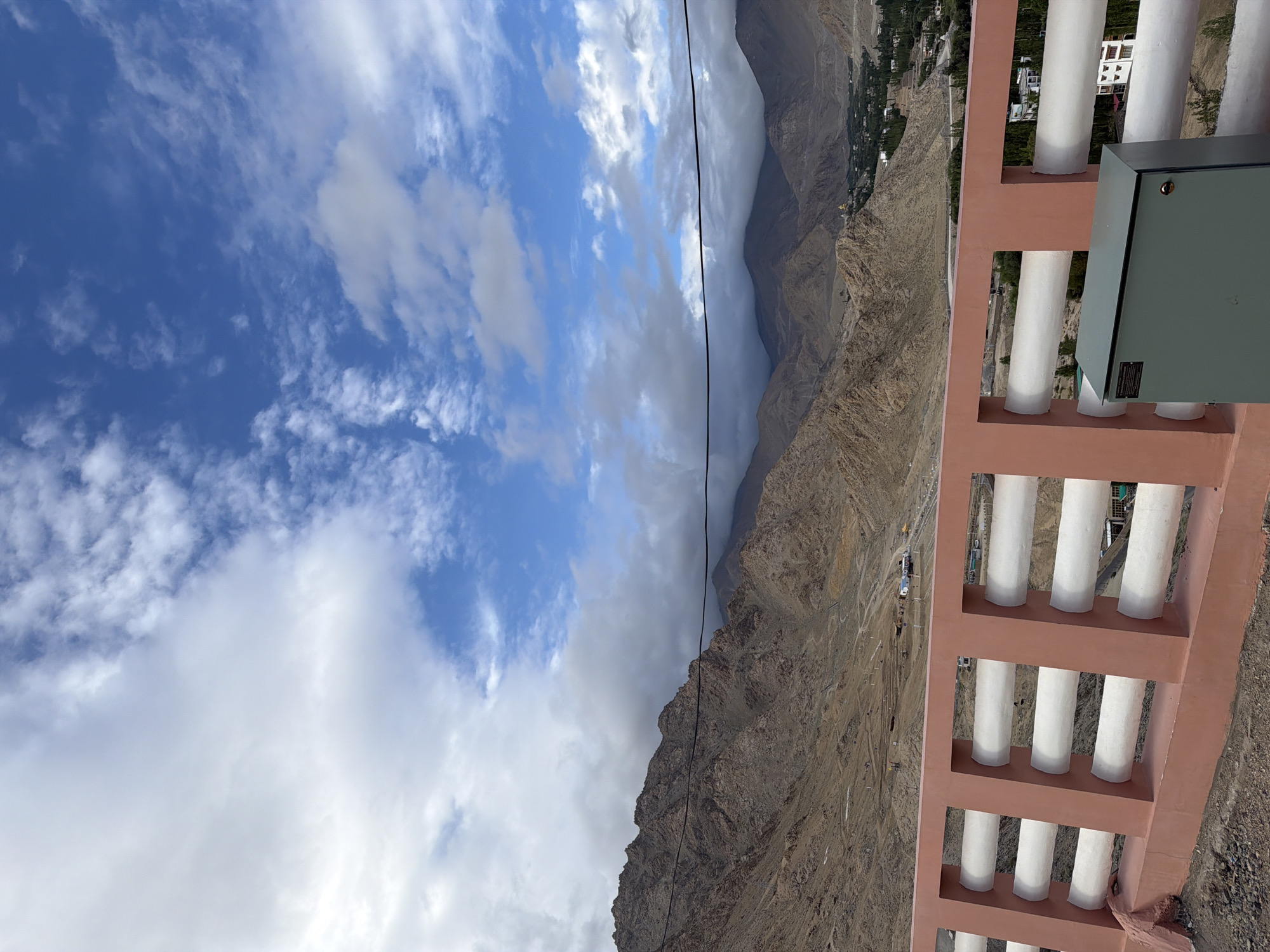 The image depicts a view from a bridge or elevated platform in Leh, India, looking out towards a mountainous landscape under a partly cloudy sky. In the foreground, a coral-colored railing with white cylindrical bars separates the viewer from the scene beyond. A light green electrical box is situated on the right side of the platform. The middle ground shows a valley with a winding road and sparse vegetation. In the background, a range of rugged, brown mountains dominates the horizon, with some peaks shrouded in clouds. A small settlement or village with some buildings and trees is visible in the far distance, nestled in the valley. The overall impression is one of a serene, arid, and mountainous environment.