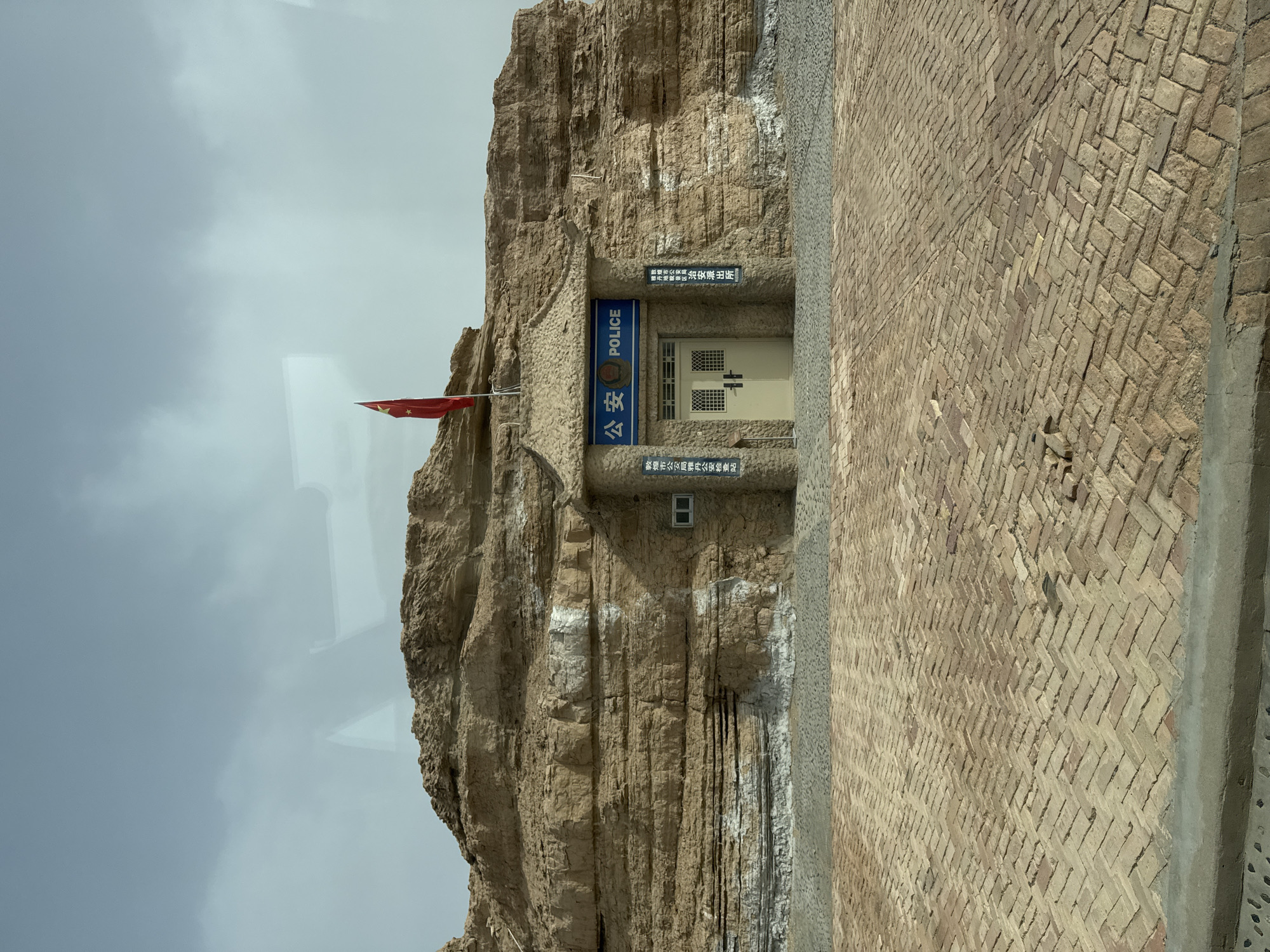 This media file captures a police station carved directly into a natural, light brown rock formation under an overcast or hazy sky. No people are visible in the scene.
The police station entrance is centrally located within the image, featuring a sturdy, light-colored double door with grid-patterned windows. Above the door, a blue sign clearly displays the Chinese characters "公安" (Gōng'ān), meaning Public Security, along with the English word "POLICE". To the left of the entrance, a Chinese national flag, red with five yellow stars, hangs from a flagpole, partly obscured.
On either side of the entrance, black vertical signs with white Chinese text are affixed to the rock. The sign on the left reads: "敦煌市公安局阳关派出所" (Dunhuang City Public Security Bureau Yangguan Police Station). The sign on the right reads: "敦煌市公安局阳关治安检查站" (Dunhuang City Public Security Bureau Yangguan Public Security Checkpoint). A small, white-framed window is visible above the right sign.
The rock face itself shows distinct layers and textures, with some white striations, suggesting weathering or mineral deposits. To the right of the rock formation, a broad, light-colored brick pathway or plaza extends into the distance, laid in a herringbone pattern. The setting suggests an arid, possibly desert, environment, consistent with Dunhuang City, China. The time of day appears to be daylight, with soft, diffused lighting indicating either an overcast sky or hazy conditions.