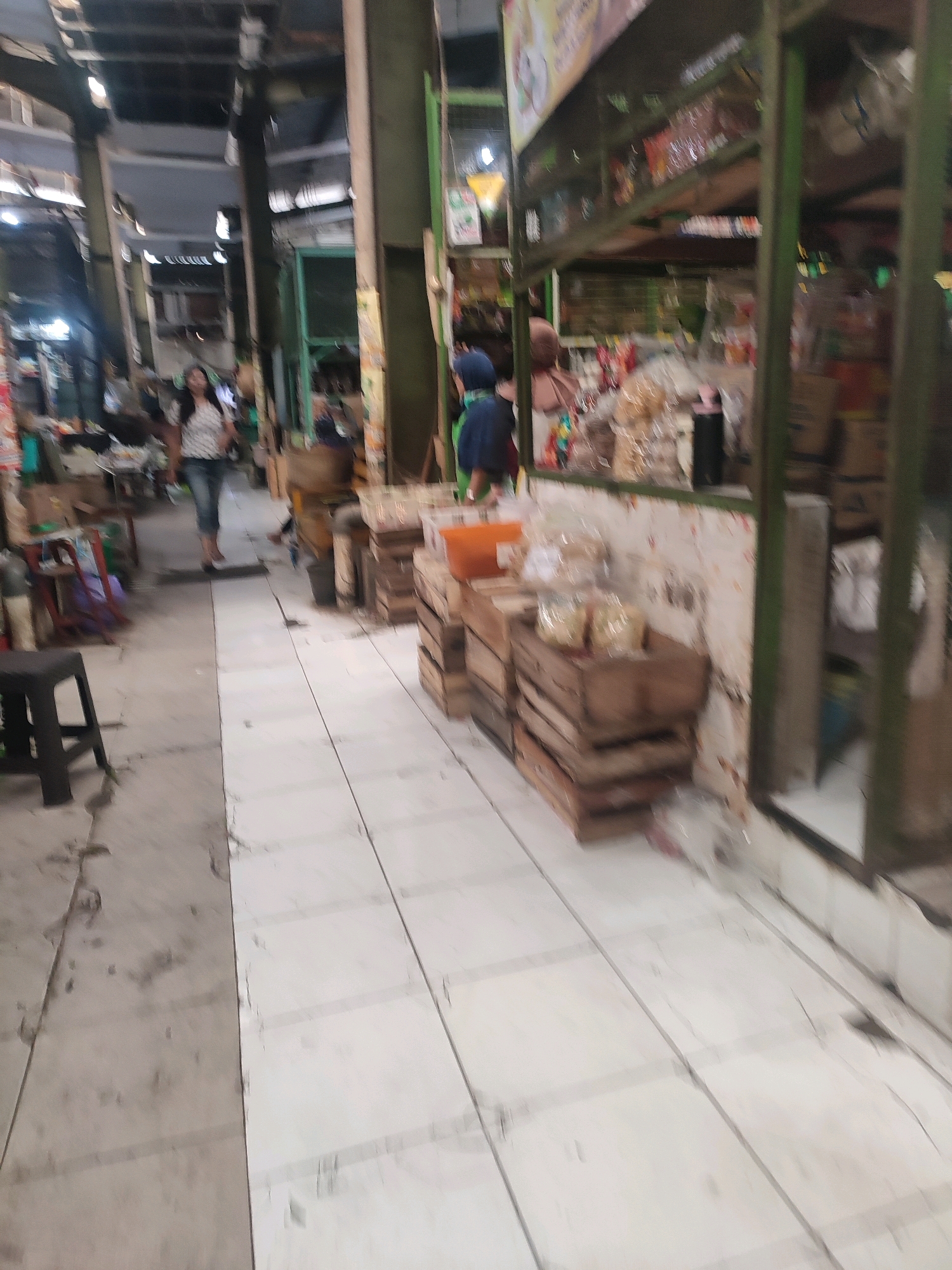 The image depicts an interior view of a market stall, likely in Yogyakarta, Indonesia. A woman in a striped shirt and jeans walks down a pathway made of white tiles. To the right of the pathway, wooden crates are stacked, some containing packaged goods. In the background, shelves are filled with various items, and other market-goers are present. The environment appears to be a busy, covered market with metal structural elements supporting the roof.