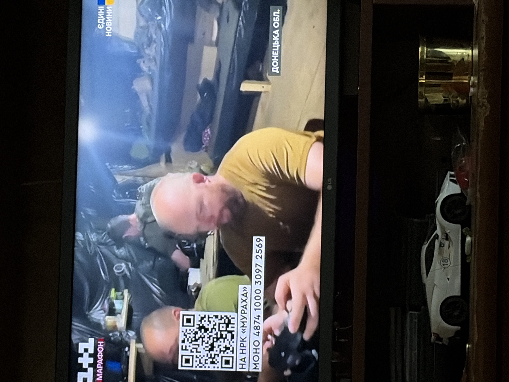 The image shows a television screen displaying a news broadcast. In the foreground of the broadcast, a QR code is visible, along with Ukrainian text that reads "НА НРК «МУРАХА» МОНО 4874 1000 3097 2569". The background of the broadcast shows two men. To the right of the screen, there is text that reads "ЄДИНІ НОВИНИ" and "ДОНЕЦЬКА ОБЛ.". Below the television screen, a white toy car with the number 18 on its side and a gold cylindrical object are visible.