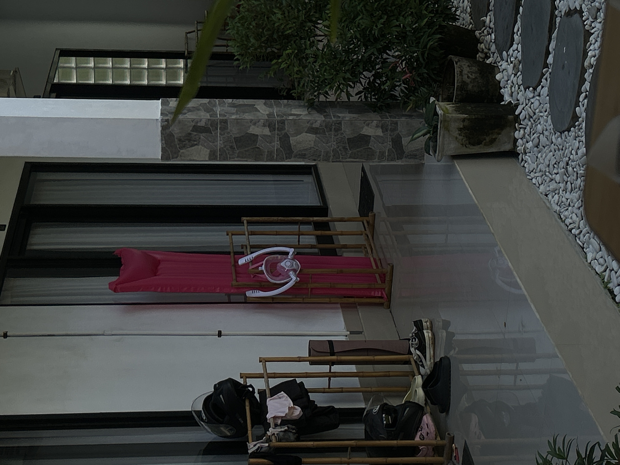 A low-angle shot captures an outdoor patio area in North Kuta, Indonesia. In the foreground, a walkway is made of dark, circular stone pavers interspersed with white gravel. To the right, potted plants are visible, one with a weathered texture. The middle ground features a tall, square pillar covered in gray stone tiles, leading to a building with a white exterior wall and large glass doors with sheer white curtains.
To the left of the pillar, a bamboo rack holds two black motorcycle helmets, one with a white stripe on top, and some clothing items. Next to this rack, a bright pink inflatable mattress leans against a similar bamboo structure. A white and clear snorkeling mask with a snorkel is attached to the mattress. Further left, more footwear is placed on the glossy, reflective floor, including black slides and white-and-black sneakers. A yoga mat is leaning against the bamboo rack. The scene is lit by soft, ambient light, suggesting it might be dusk or early morning.