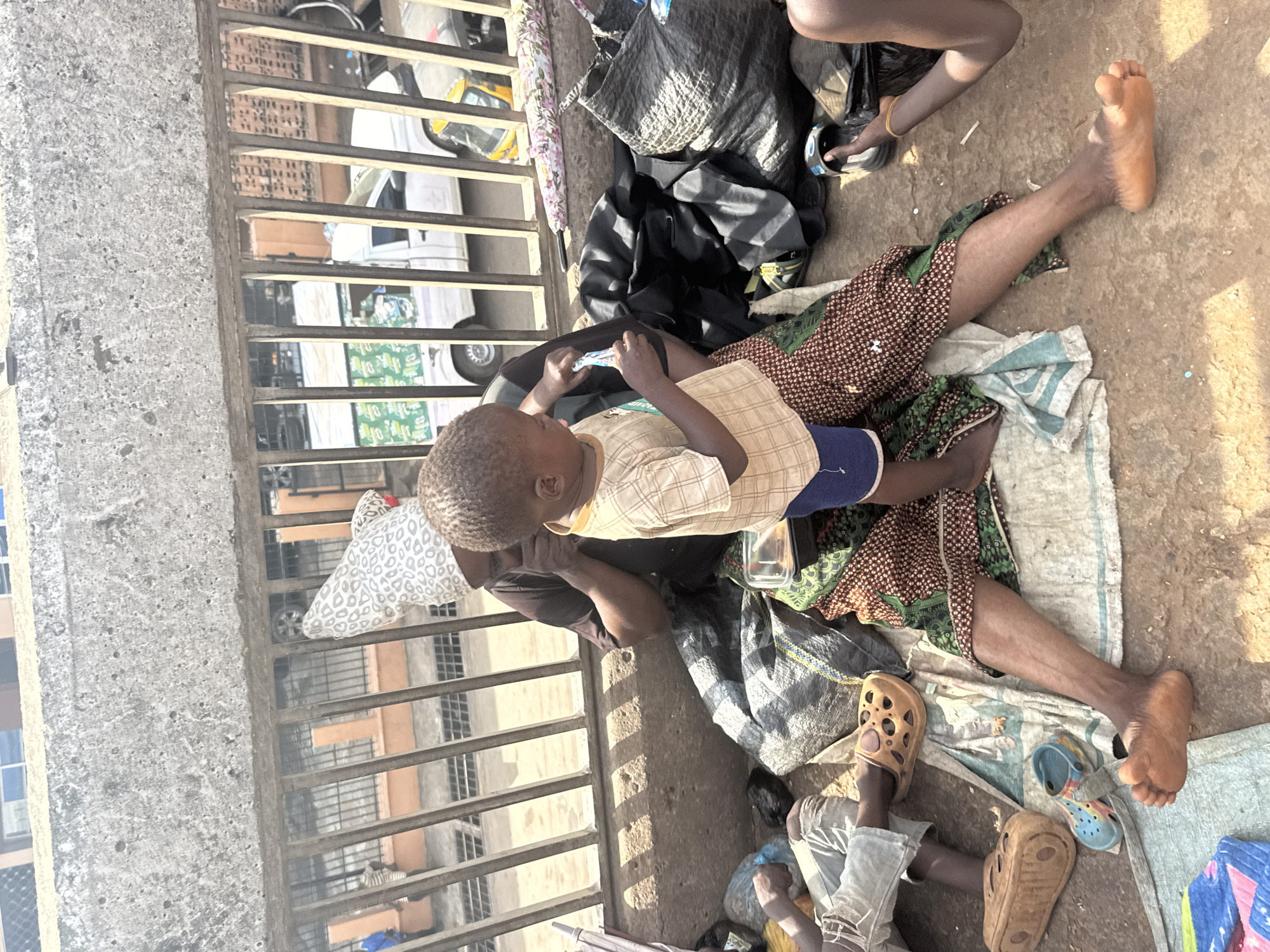 A person with dark skin lies prone on a section of concrete in a public space, their legs splayed. They are wearing a patterned wrap skirt, and a child in a plaid shirt and dark shorts lies on top of them. A second child sits nearby, wearing light-colored pants and brown sandals. Luggage and bags are scattered around the person and children. A metal fence is situated to the left, with the city street visible in the background. The scene is located in Onitsha, Nigeria.