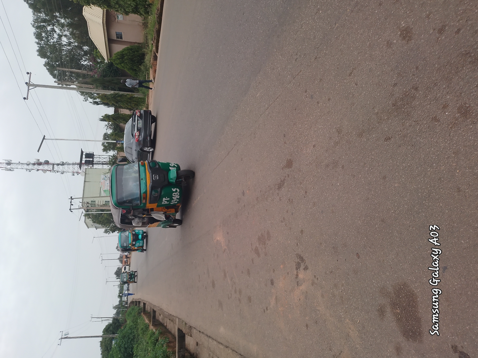 A street scene in Jos, Nigeria features a prominent green and yellow auto-rickshaw, or "Keke Napep," driving on a paved road. The Keke Napep has the license plate "PT-19485" and appears to have a driver and passengers. Other vehicles, including a dark-colored sedan and additional Keke Napeps, are visible in the background, along with utility poles, trees, and buildings. The sky is overcast. The foreground displays a section of the asphalt road, marked with the text "Samsung Galaxy A03."