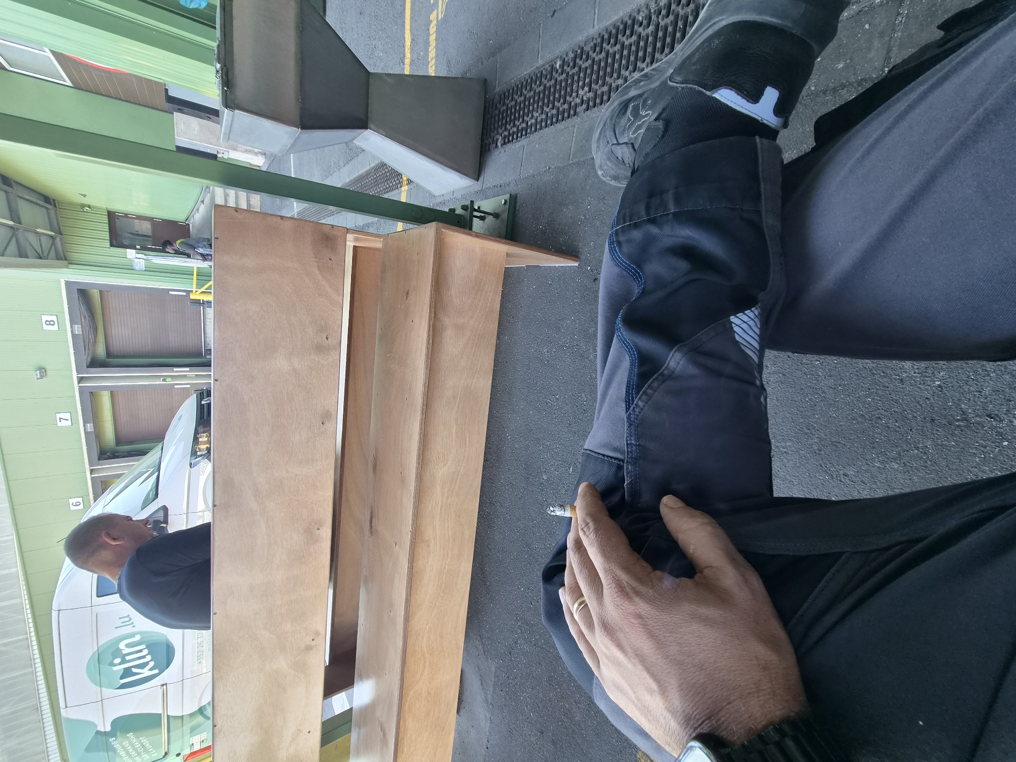 An individual, seen from the waist down, is seated on a wooden bench with a lit cigarette in their right hand. The person is wearing dark pants and black shoes. The background features a white van with "klin.lu" branding, a green building with loading bays numbered 6, 7, and 8, and a concrete structure. The scene is outdoors, on asphalt, suggesting an industrial or commercial area. The van's branding indicates the location is likely within or near Leudelange, Luxembourg, where "klin.lu" operates as a laundry service.