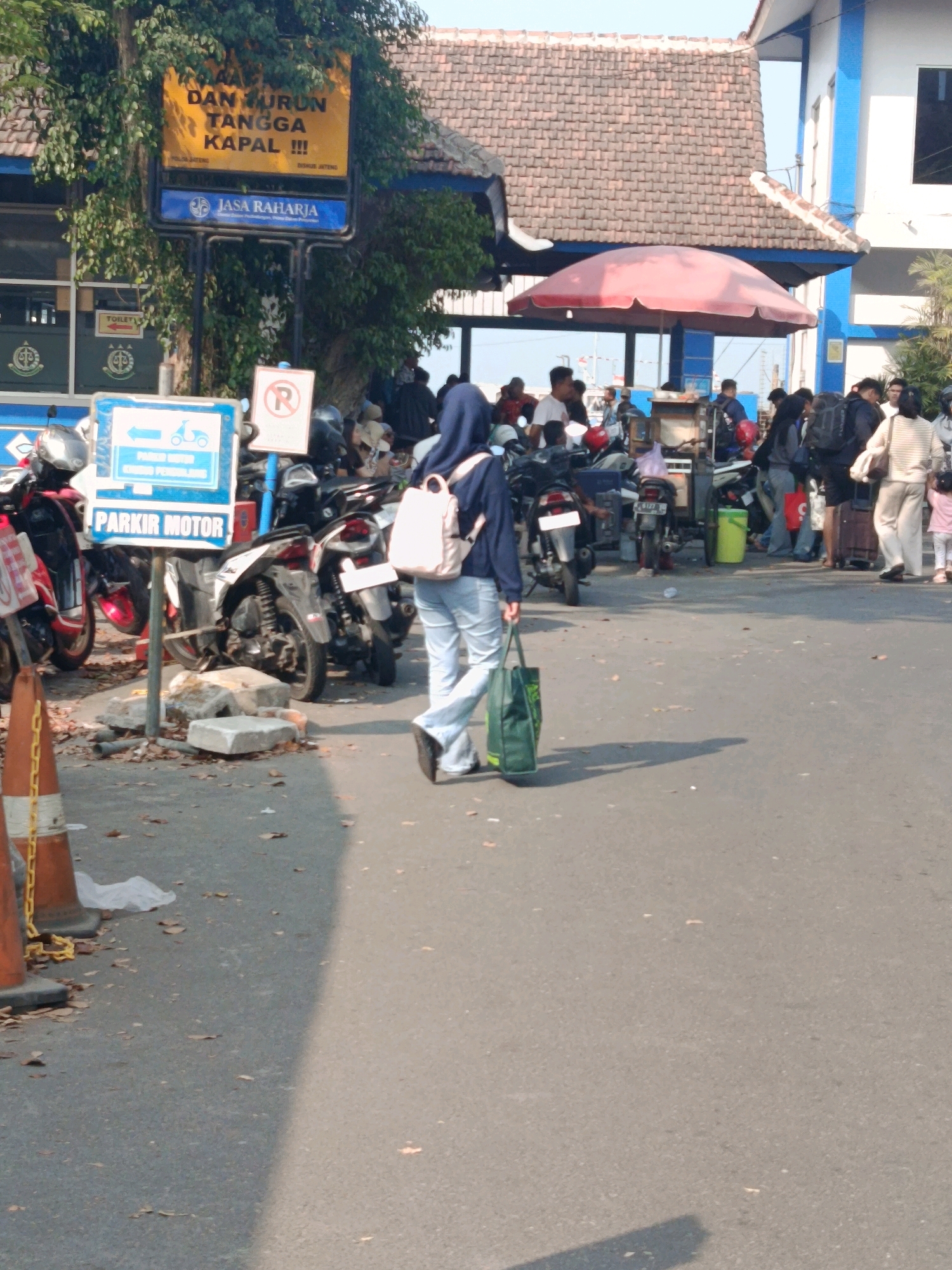 A woman wearing a navy blue hijab and light blue jeans walks away from the camera, carrying a green tote bag in her right hand and a light-colored backpack on her shoulders. She is walking on an asphalt road in what appears to be a port or ferry terminal. To her left, there are several parked motorcycles, one of which has a blue and white sign with a motorcycle icon and the text "PARKIR MOTOR" (Motorcycle Parking). Behind the woman, there is a crowd of people, some with luggage, and more motorcycles. In the background, there is a building with a tiled roof and blue trim, and a large yellow sign with Indonesian text. The scene is outdoors, with sunlight casting shadows on the ground. The overall impression is of a busy transit point.