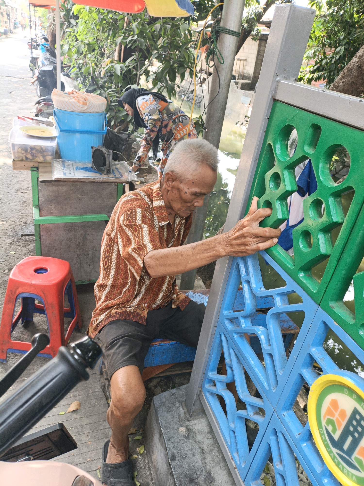 In the city of West Jakarta, Indonesia, an elderly man with grey hair and a patterned shirt sits by a roadside stall. He is wearing dark shorts and black sandals, and his right hand rests on a decorative blue and green barrier. Behind him, a woman wearing a headscarf and a colorful patterned top is visible, tending to the stall. The stall displays various items, including blue plastic buckets, containers with food, and what appears to be a speaker. A red plastic stool is positioned to the left of the man. The environment suggests an outdoor setting with trees and a glimpse of water or a canal in the background.