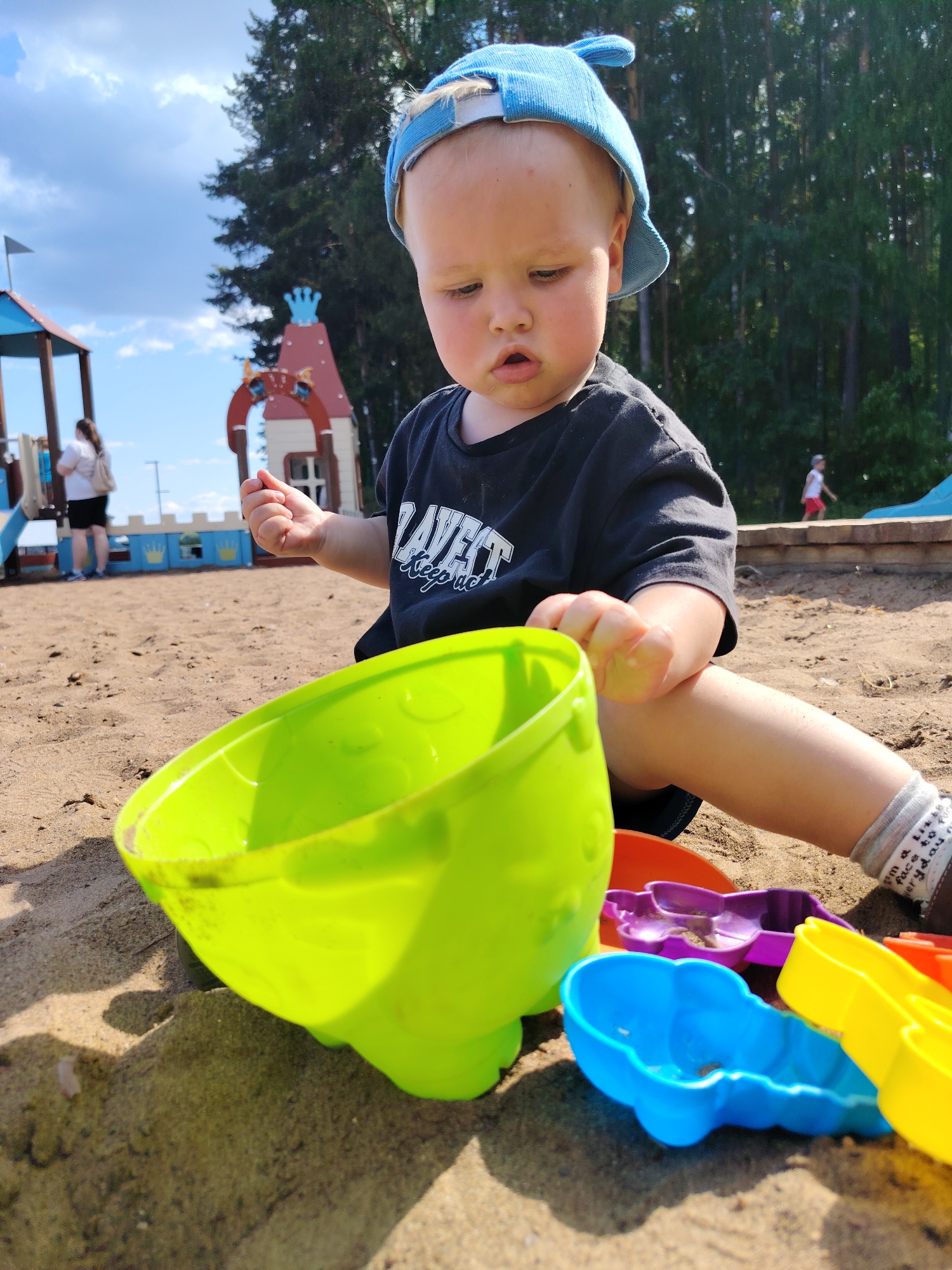 The image shows a young child in a sandbox, playing with colorful plastic toys. The child, wearing a blue cap and a black t-shirt, is seated in the sand, holding a small object in their hand and looking down at a large green bucket. Several brightly colored sand molds are visible in the foreground, including blue, purple, and yellow ones. In the background, there is a playground structure with a red and white castle-like design and a blue slide. Two adults are also present in the background, one standing near the playground structure and another walking further away. The scene is set outdoors on a sunny day with a cloudy sky, and trees are visible in the background. The location is identified as Chaikovsky, Russia.