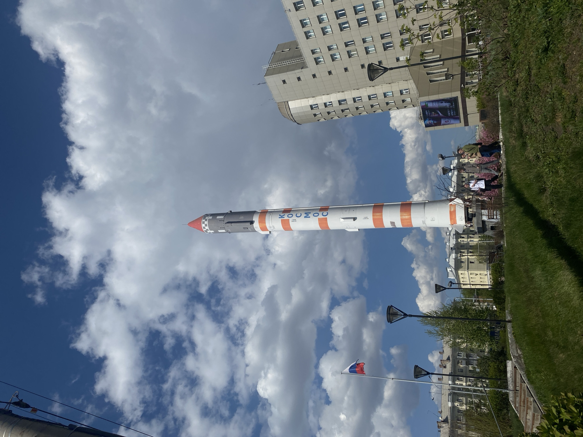 Here is a concise description of the image:
The image shows a horizontal view of a rocket monument, identifiable as a Soyuz rocket, set against a partly cloudy sky. The text "COЮ3 MO" (Soyuz) is visible on the side of the rocket. In the background, a large modern building with multiple windows is partially visible. A park-like area with green grass and trees is to the right of the rocket. Street lamps are lined up nearby, and a Russian flag is visible on a pole. People appear to be walking around the monument. The scene suggests the rocket is a public display or memorial in Krasnoyarsk, Russia.