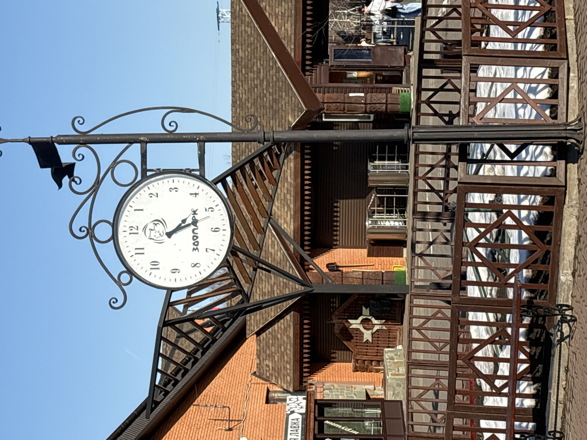 The image showcases a street clock in Izhevsk, Russia, with the clock face displaying the time as approximately 4:57. The clock is mounted on a decorative iron post, with an elaborate design, and it has the inscription "300ПАРК" (300Park) on its face, alongside a stylized animal image. The background consists of a multi-story building with architectural details, including decorative woodwork, brickwork, and windows. A sign with Cyrillic text (ПЛАВКА) is visible near the bottom of the frame. The sky is clear and blue, indicating a sunny day.