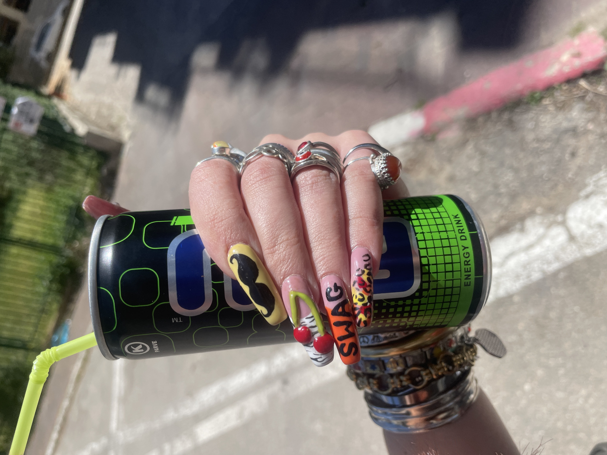 A hand is holding a black and green energy drink can with a yellow straw, with elaborate nail art on each finger, including a mustache, cherries, "SWAG" text, and leopard print. The hand is adorned with multiple rings on each finger and bracelets on the wrist. The scene appears to be outdoors, possibly on a sidewalk or street, with a building and greenery visible in the background. The exact location in Raanana, Israel cannot be identified without additional context.