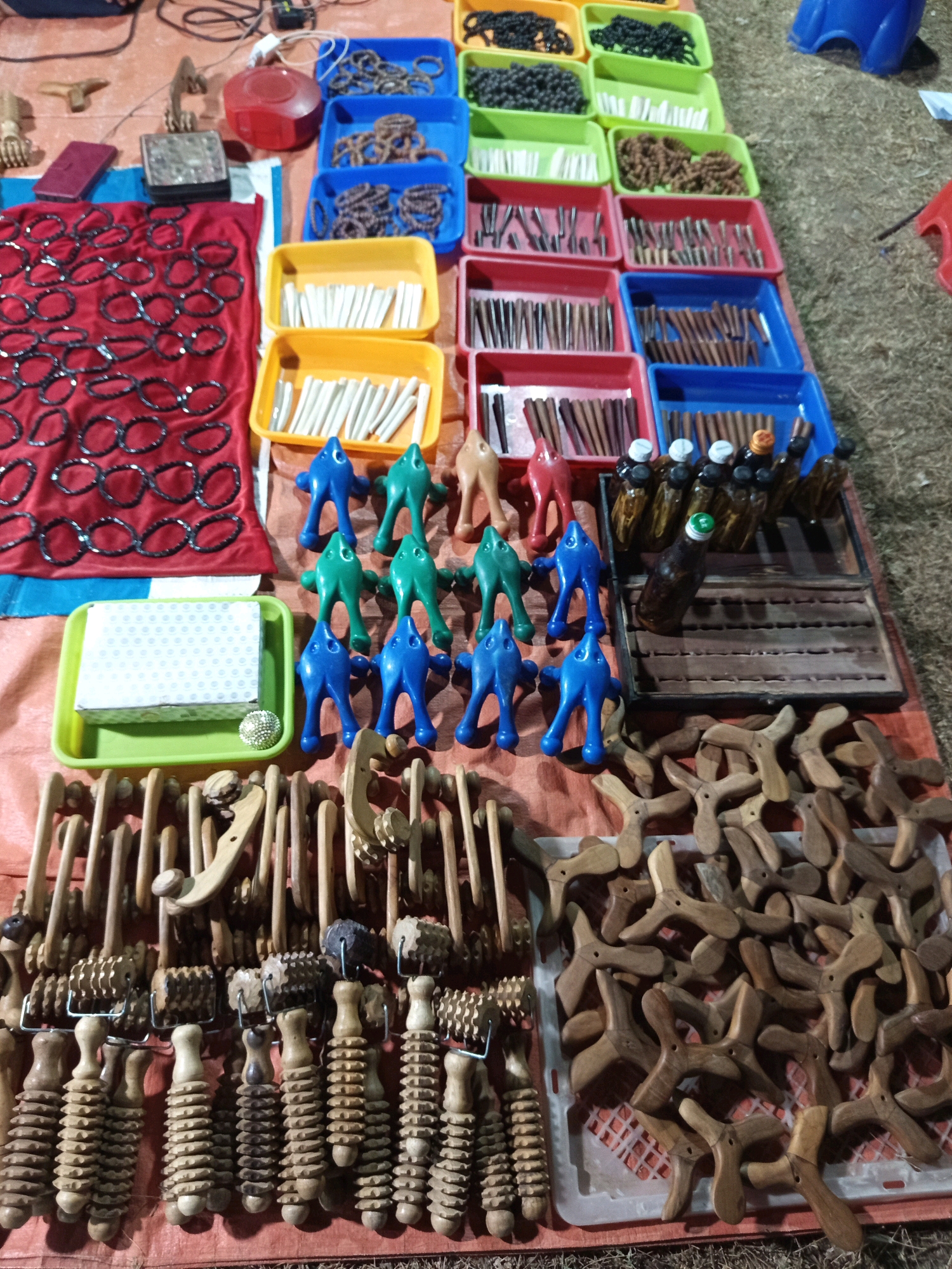 The image depicts an outdoor market stall, likely in Pulung Kencana, Indonesia, displaying a variety of handcrafted items for sale. The stall is set up on a textured, orange-brown tarp laid over a grassy or dirt surface. Various containers, primarily plastic trays in blue, green, and yellow, are arranged in rows, holding different types of merchandise.
Prominently featured are numerous wooden massage tools, including rollers with textured surfaces and smaller, handheld massagers shaped like animals (blue and green figures) and abstract wooden pieces, some resembling star shapes or tripods. There are also bundles of small wooden sticks, possibly incense or massage aids, neatly arranged in red and blue trays. In one section, a red cloth is spread out, covered with black bracelets or necklaces.
Several small glass bottles, some containing a dark liquid and others appearing empty, are arranged in a wooden display rack. A red plastic container is also visible. The lighting suggests it might be evening or a dimly lit indoor setting, with some items casting shadows. No people are directly visible in the frame, but the arrangement implies a commercial transaction setting. The overall scene is one of organized retail display of artisanal products.