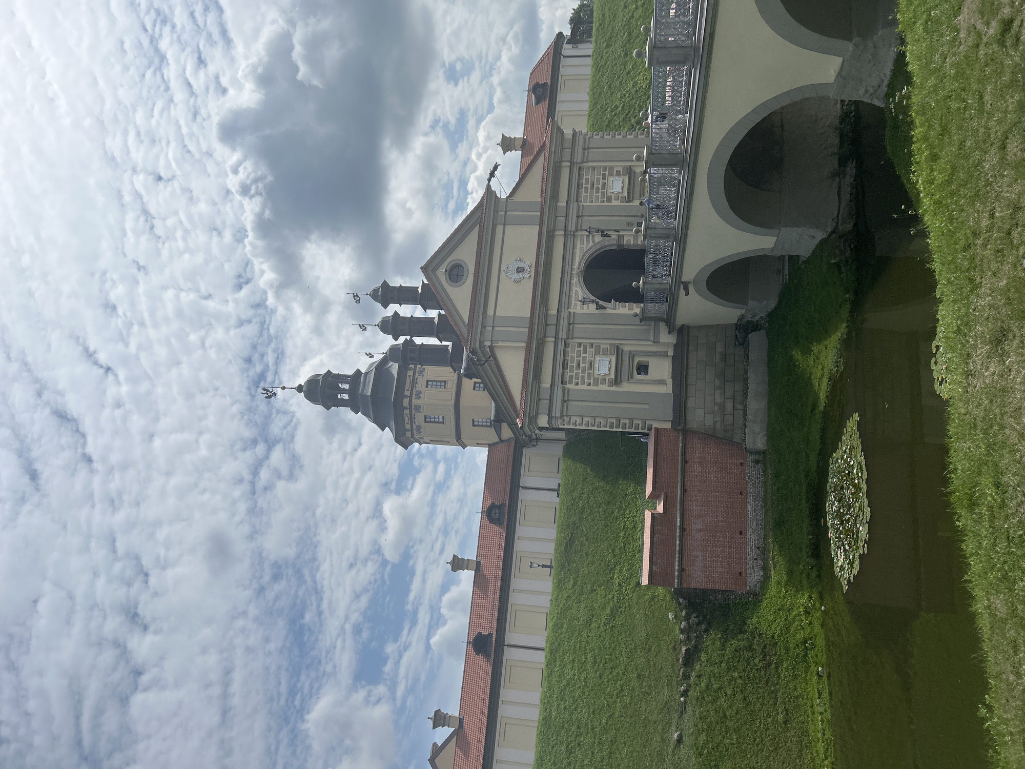 The image depicts a view of Nesvizh Castle, a UNESCO World Heritage site, in Nesvizh, Belarus. The castle complex features a prominent gatehouse with arched entranceways, flanked by towers and connected to the main structure by a bridge with ornate railings. The buildings are characterized by red tiled roofs and pale yellow walls, with architectural details like decorative crests and dormer windows.
The castle is situated on a grassy hill, with a moat or pond in the foreground containing lily pads. A brick wall is visible on the lower left side of the image. The sky above is partly cloudy, with a mix of white clouds against a blue sky. A few individuals can be seen on the bridge, suggesting a public accessible area. The overall impression is one of historical architecture and natural scenery.