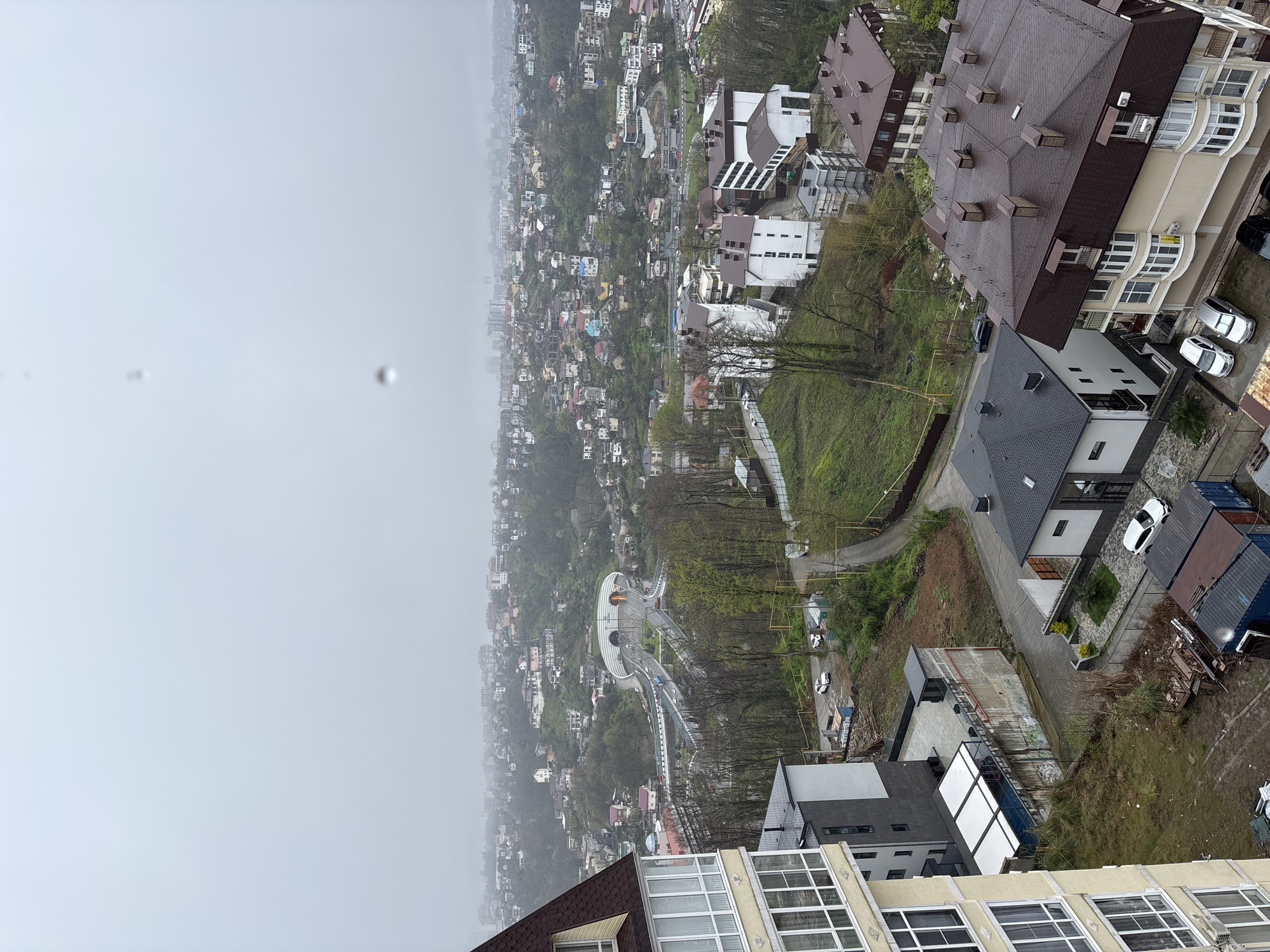 This image depicts a cityscape in Sochi, Russia, shrouded in fog. The view is taken from a high vantage point, likely from atop a tall building. In the foreground are buildings with varied architectural styles, featuring details such as brown and gray roofs, balconies, and large windows. A few cars are parked near these buildings.
Beyond the immediate foreground, the city stretches out into the distance, with dense clusters of houses and buildings nestled amongst greenery. The overall appearance suggests a hilly or mountainous terrain. A distinctive feature in the mid-ground is a white, winding road or walkway structure, possibly a bridge or elevated path, with a tunnel-like feature along its course.
The environment is overcast and foggy, obscuring distant details and giving the scene a muted color palette. The lack of clear definition indicates conditions of reduced visibility. No people are immediately apparent in the view.